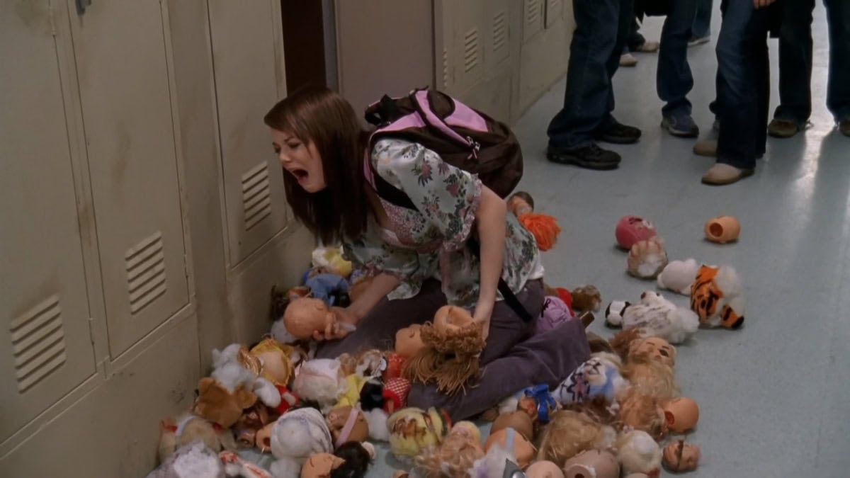 Emma Stone, as Diane, wears a floral blouse with lavender corduroy pants, a pink-trimmed backpack, and brown closed-toe wedge clogs—an unmistakably mid-2000s look that contrasts humorously with her character’s mean-girl antics in the episode