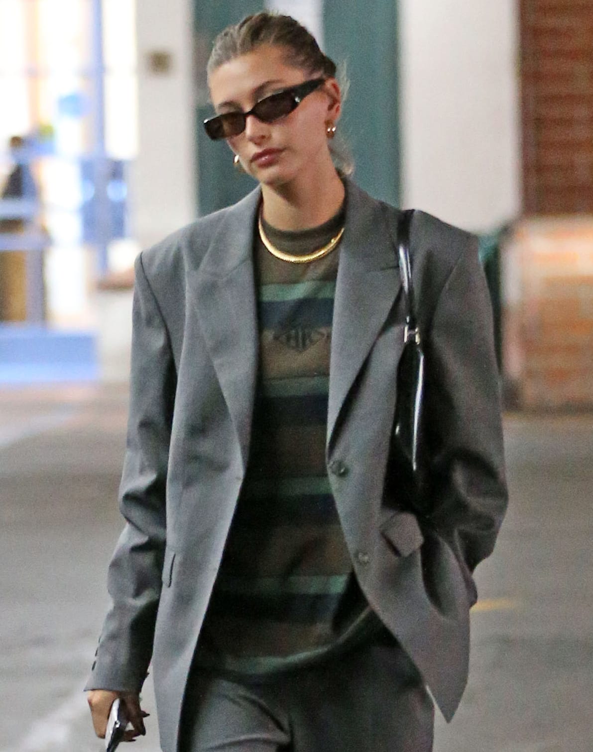 Hailey Bieber wears a messy bun and styles her masculine outfit with gold jewelry, Raen Tongue sunglasses, and a Prada bag