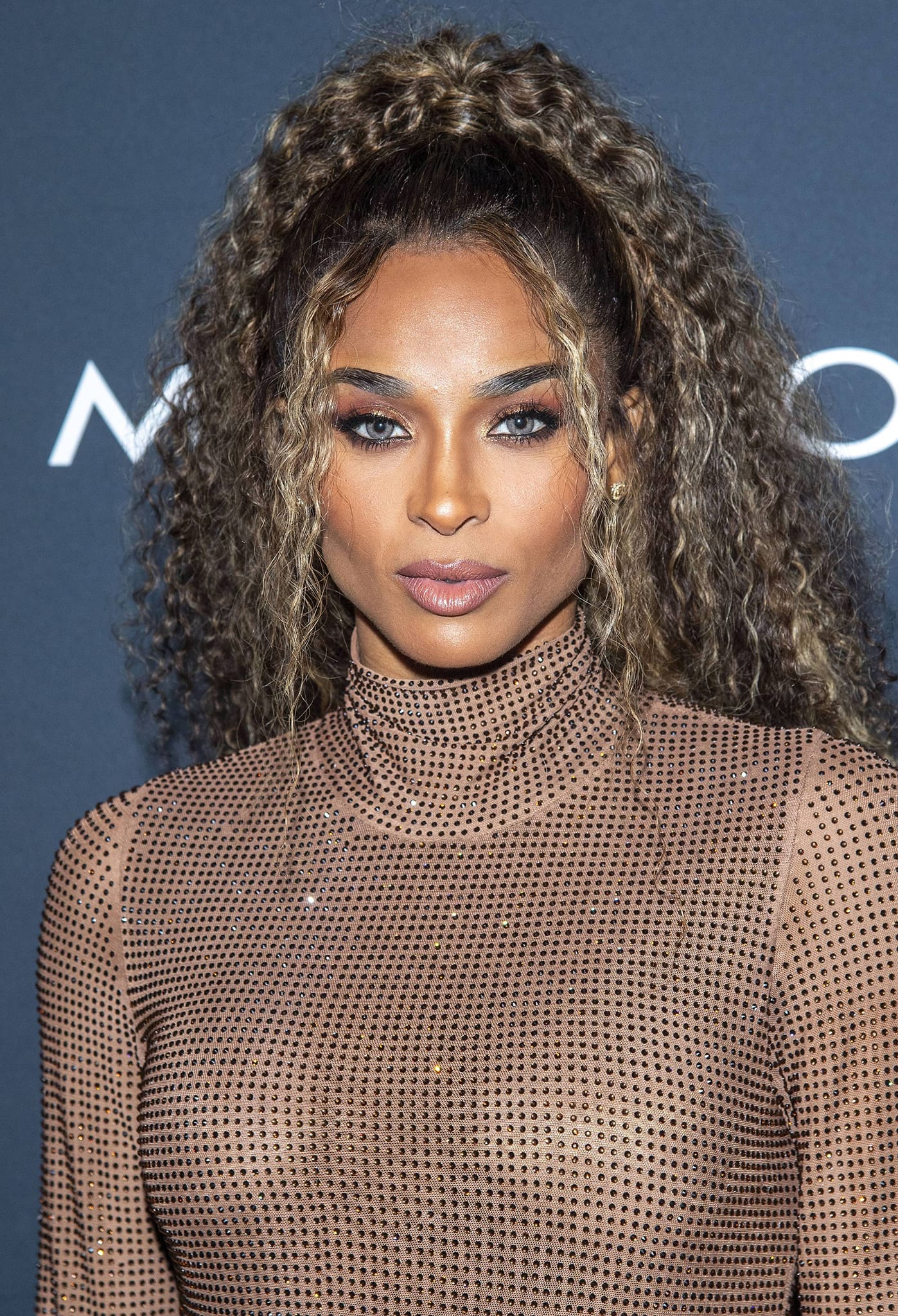 Ciara glams up with a curly ponytail wig, smokey eyes, hazel lenses, and nude pink lipstick