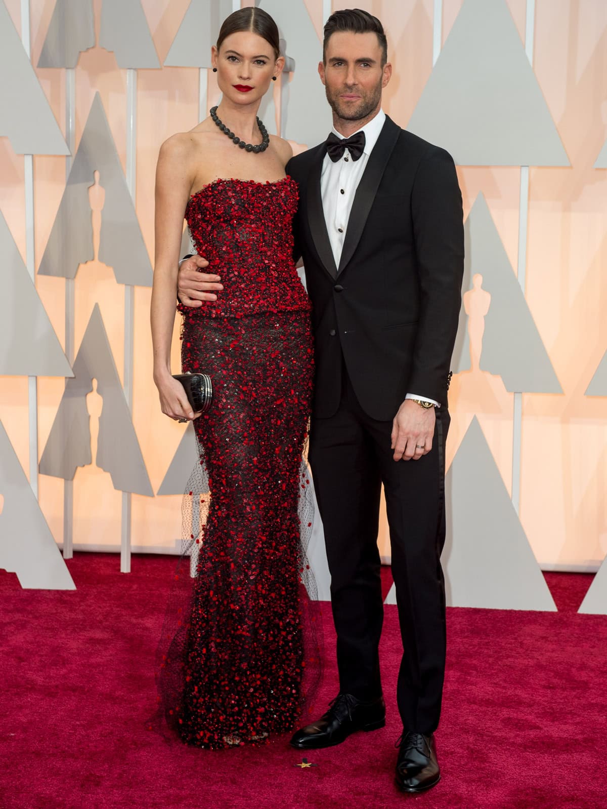 Behati Prinsloo stuns in a strapless black-and-red beaded Armani Privé gown, elevating the sleek column silhouette with hidden high-heeled sandals as she poses with Adam Levine at the 87th Annual Academy Awards on February 22, 2015