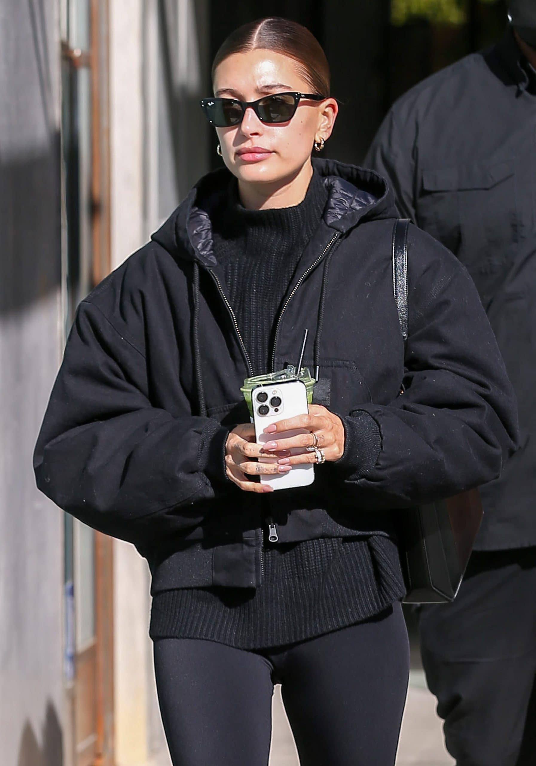 Hailey Bieber wears her signature center-parted tight bun with minimal makeup and RayBan angular sunglasses