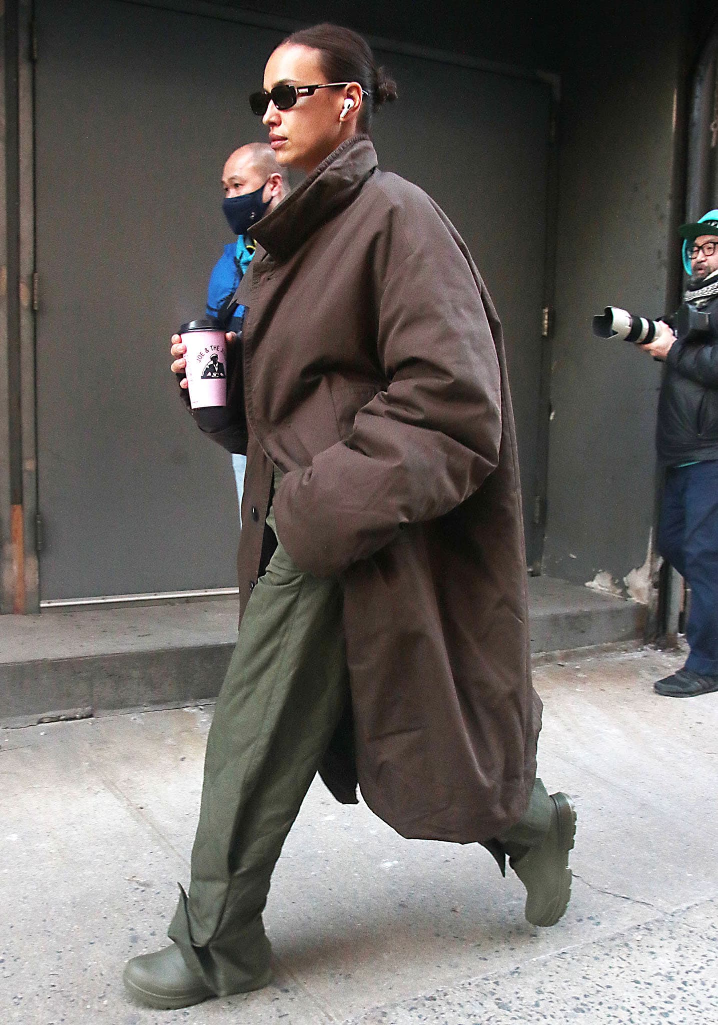 Irina Shayk grabbing a cup of coffee in tonal beige crop top, loose green pants, and brown coat