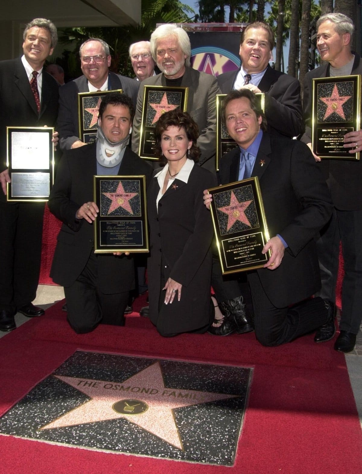 Donny, Marie, and Jimmy Osmond are seated with Jay, Tom, family patriarch George, singer Andy Williams, Merrill, Allan, and Wayne Osmond standing at a ceremony honoring The Osmond family with a star on the Hollywood Walk of Fame