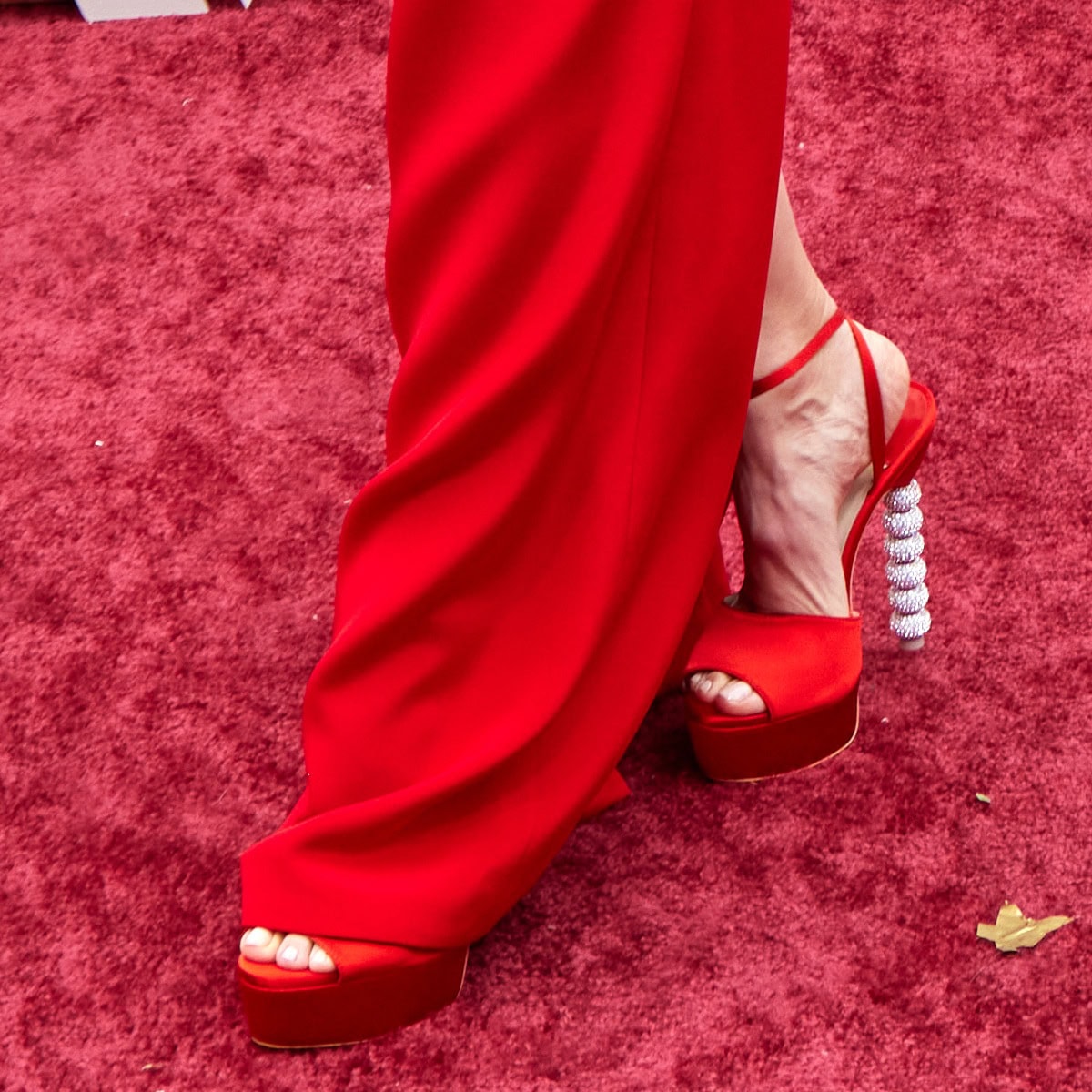 Sophia Webster’s red platform sandals add dramatic height and sparkle, their crystal-embellished heels perfectly echoing the bold glamour of the flowing scarlet gown on the Oscars red carpet