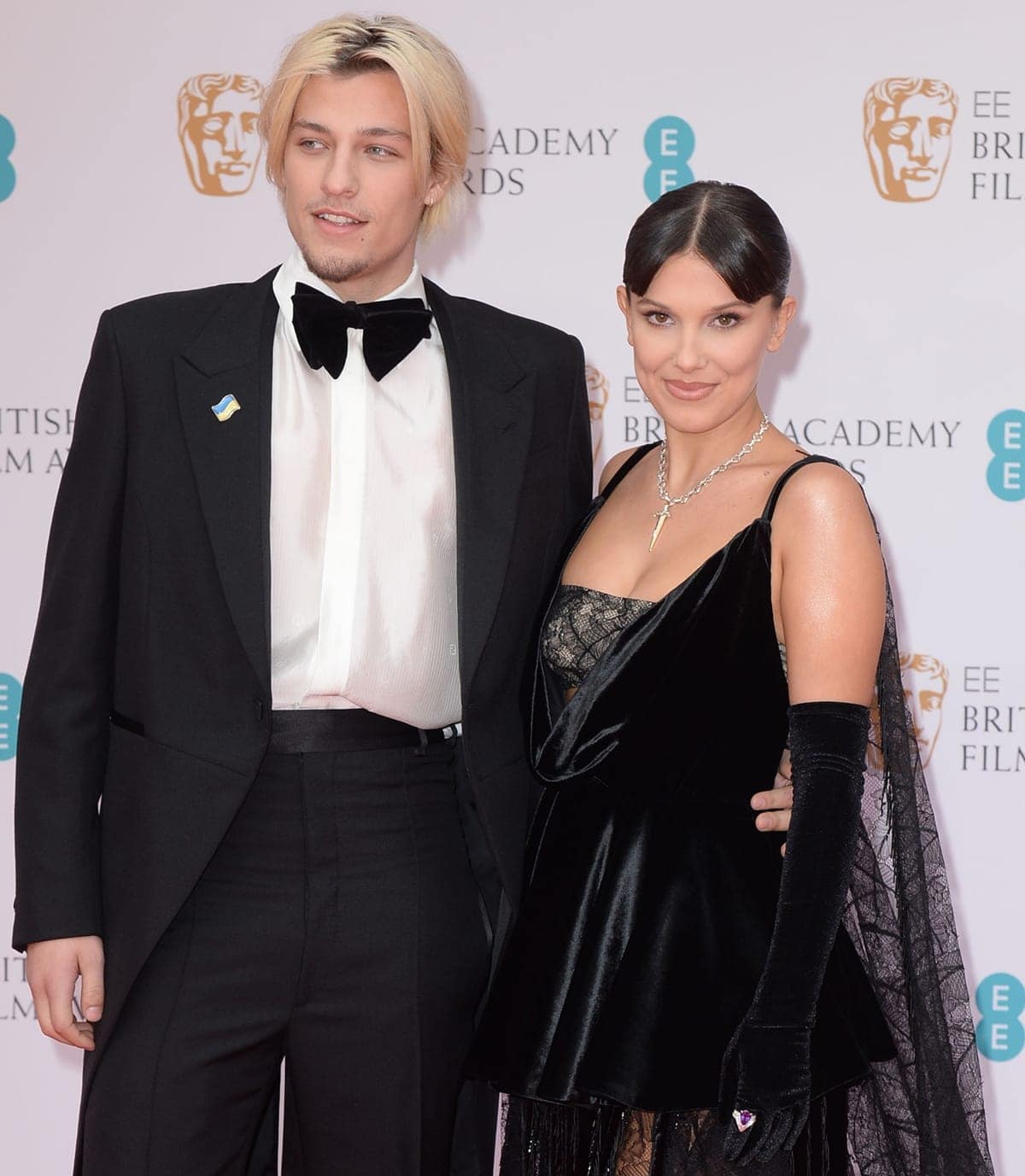 Millie Bobby Brown and Jake Bongiovi make a striking pair at the BAFTAs, with Brown in a black velvet–and–lace Louis Vuitton dress and opera gloves while Bongiovi complements her in a classic tux accented by a Ukrainian flag pin