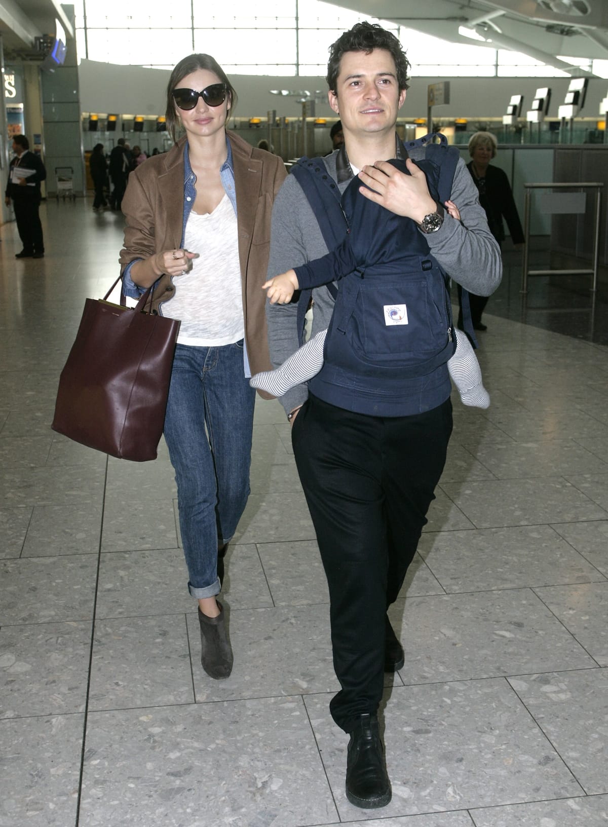 Miranda Kerr keeps it effortlessly chic at Heathrow Airport in cuffed skinny jeans, suede ankle boots, and a casual blazer, while Orlando Bloom stays comfy in sneakers and a baby carrier for their son, Flynn