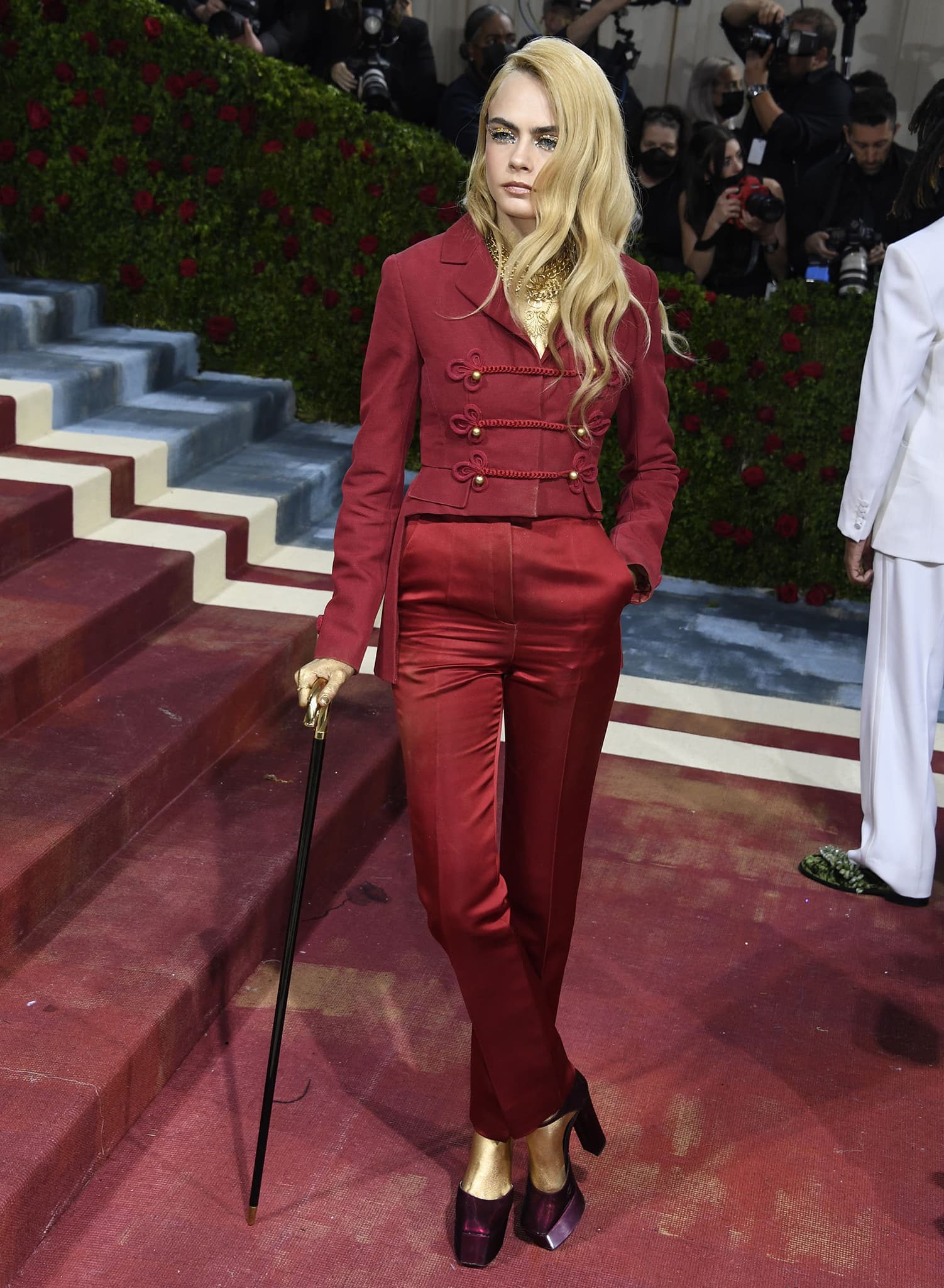 Cara Delevingne arrives on the red carpet in a Dior red suit, burgundy platform heels, and a gold cane