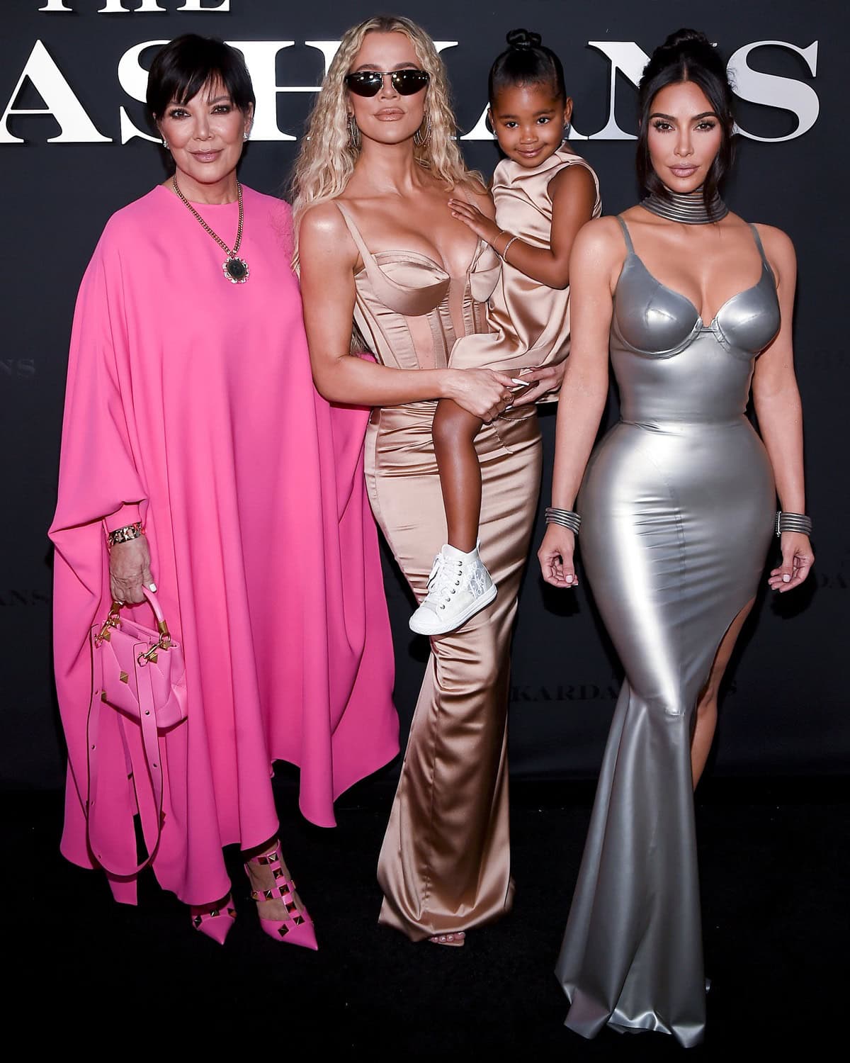 Kris Jenner wears pink studded heels, Khloé Kardashian holds her daughter True Thompson who is wearing white sneakers, and Kim Kardashian's shoes are not visible