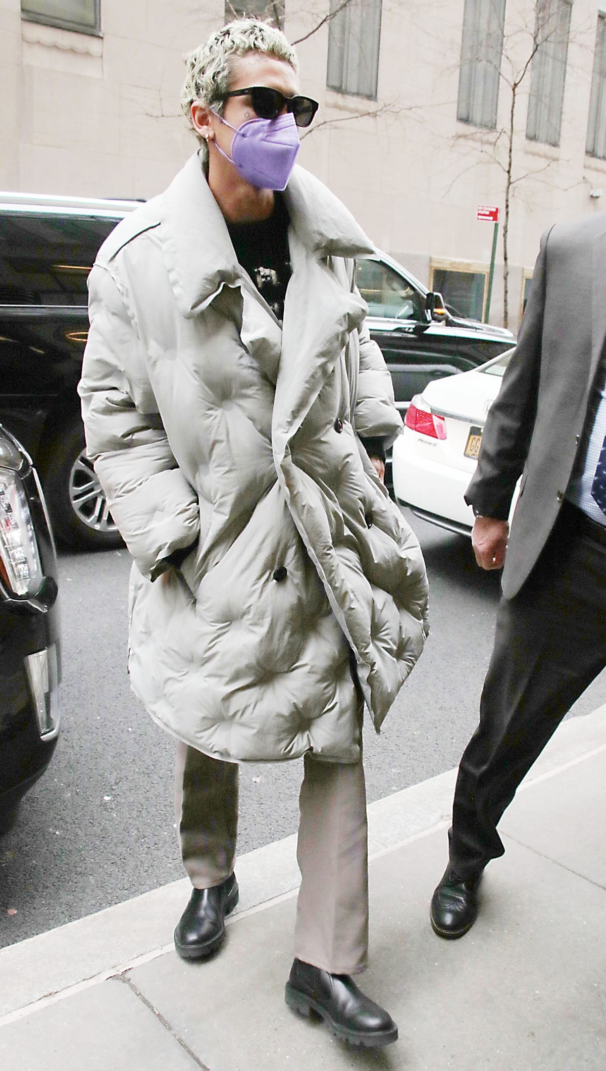 Dominic Fike wears a purple face mask and a light gray long puffer coat in New York City