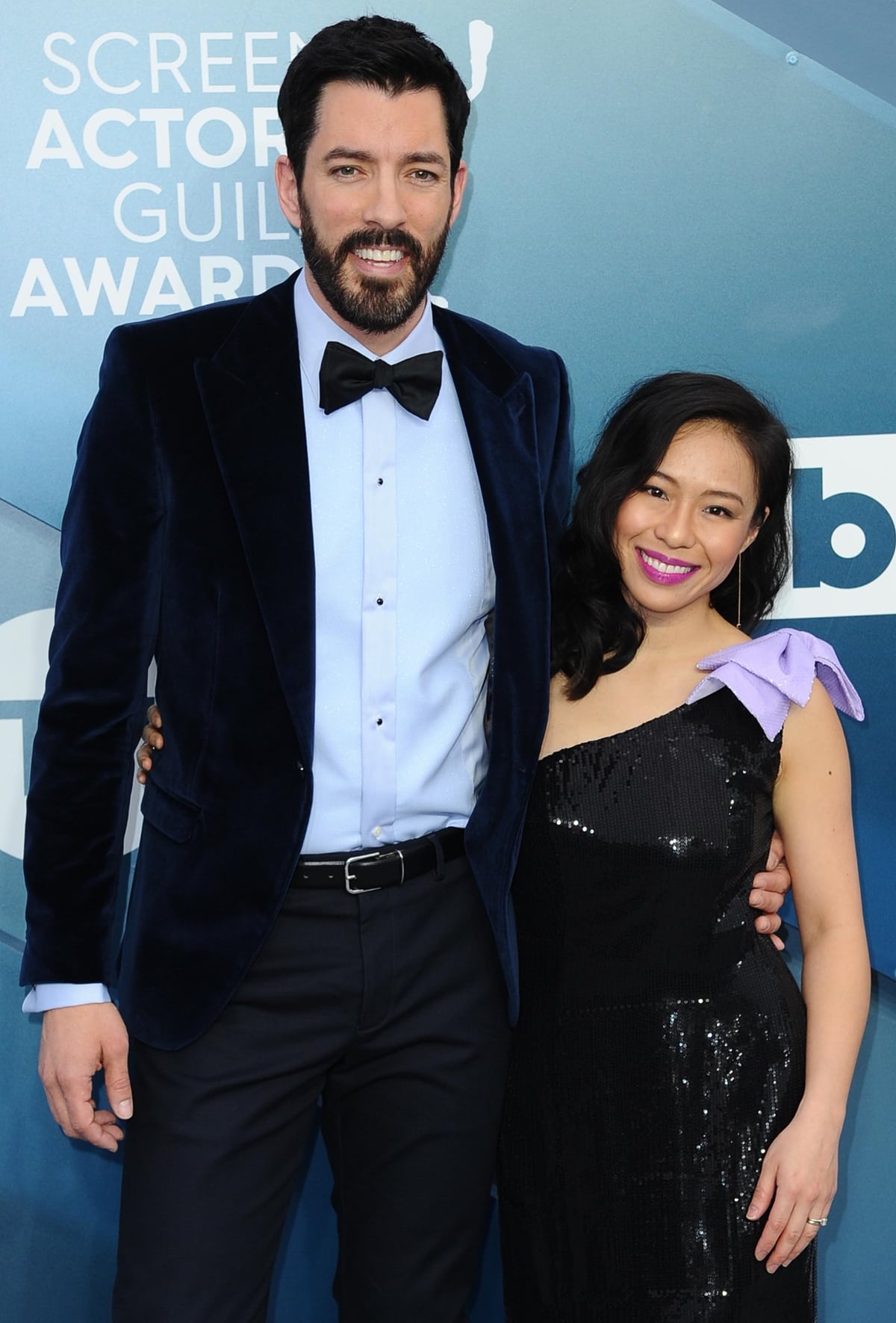 Drew Scott exudes classic charm in a velvet tuxedo while Linda Phan stuns in a black sequin one-shoulder gown with a playful lavender bow accent