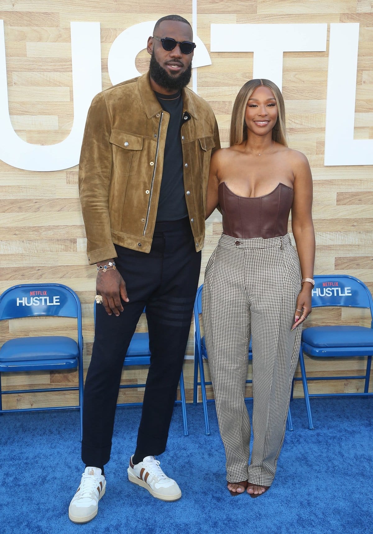 LeBron James and Savannah James pose in stylish outfits at the Netflix world premiere of Hustle—LeBron in a suede jacket and sneakers, while Savannah stuns in a brown leather bustier and checkered trousers
