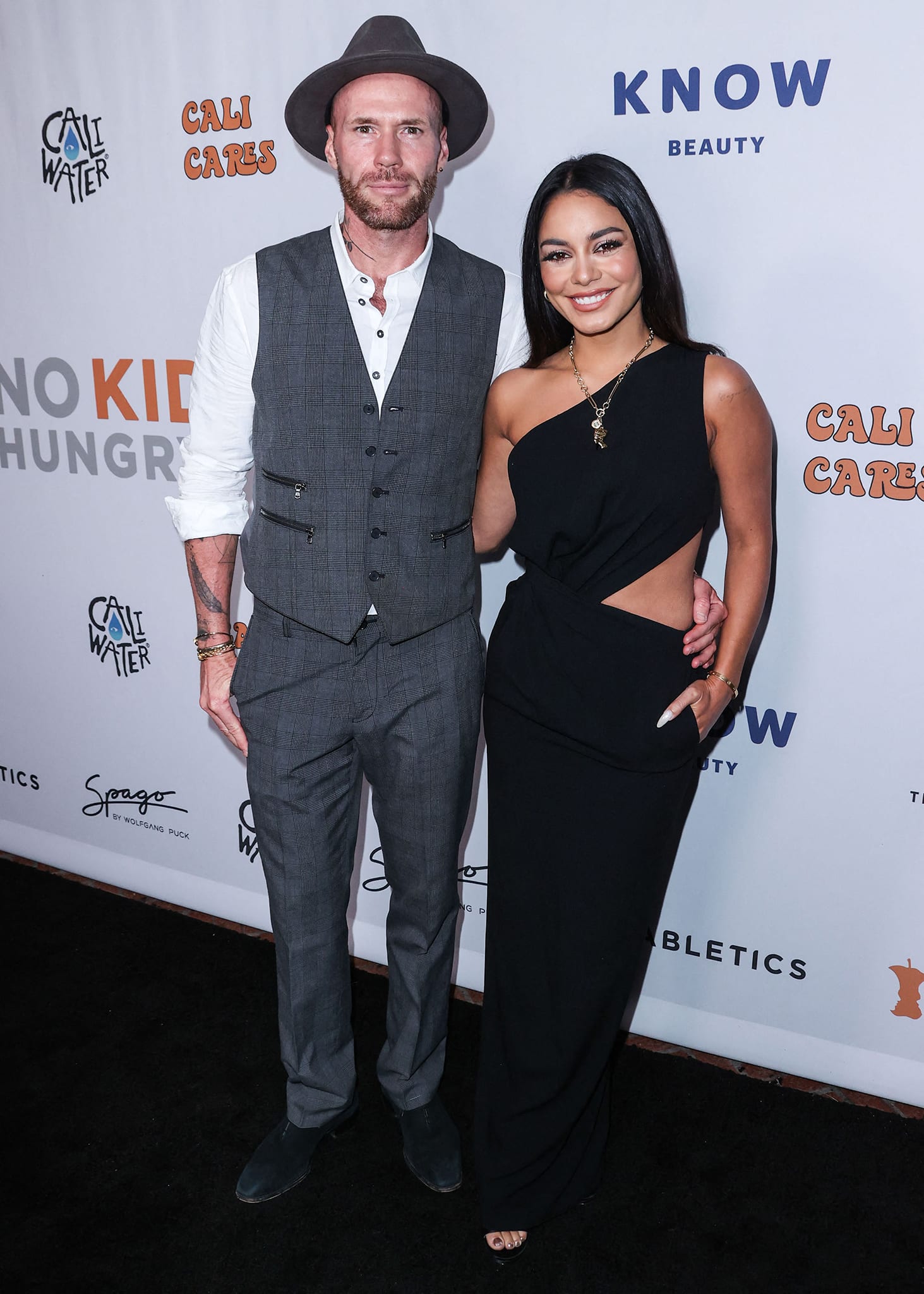 Caliwater co-founder Oliver Trevena joins Vanessa Hudgens on the black carpet in a patterned gray vest, white shirt, and a hat