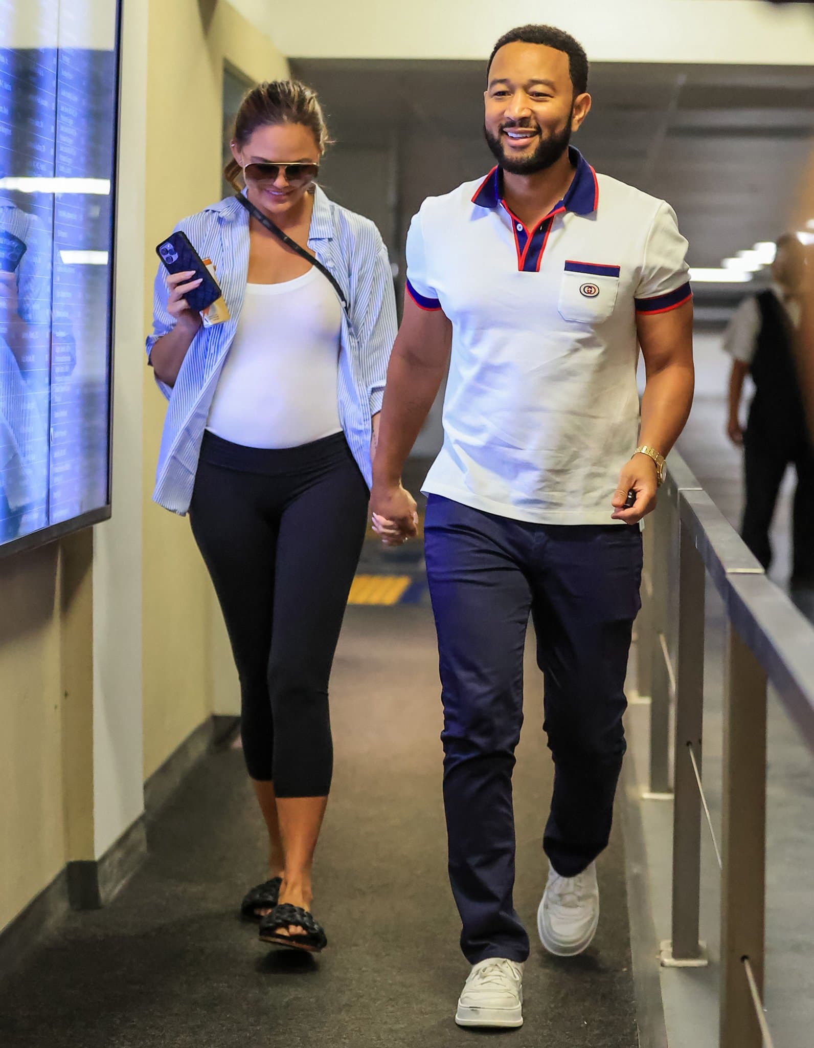 Pregnant Chrissy Teigen shows off her growing baby bump in a fitted white top, black leggings, and unbuttoned light blue shirt