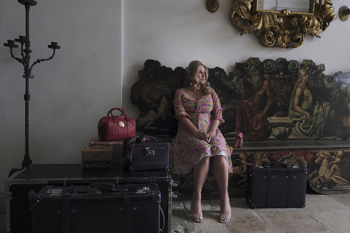 Jennifer Coolidge as Tanya McQuoid-Hunt in The White Lotus Season 2, sitting among her designer luggage in Sicily while wearing a floral Emanuel Ungaro dress and clear Betsey Johnson PVC block-heel sandals—a costume choice that later became central to her viral finale scene