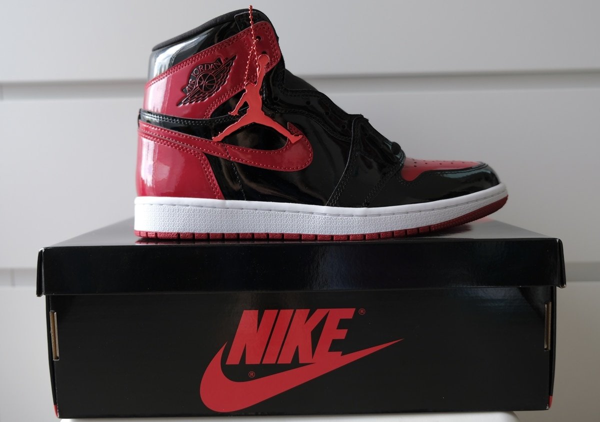 The Air Jordan 1 High Bred Patent features a black and red patent leather upper with signature weaved Nike Air tongue labels