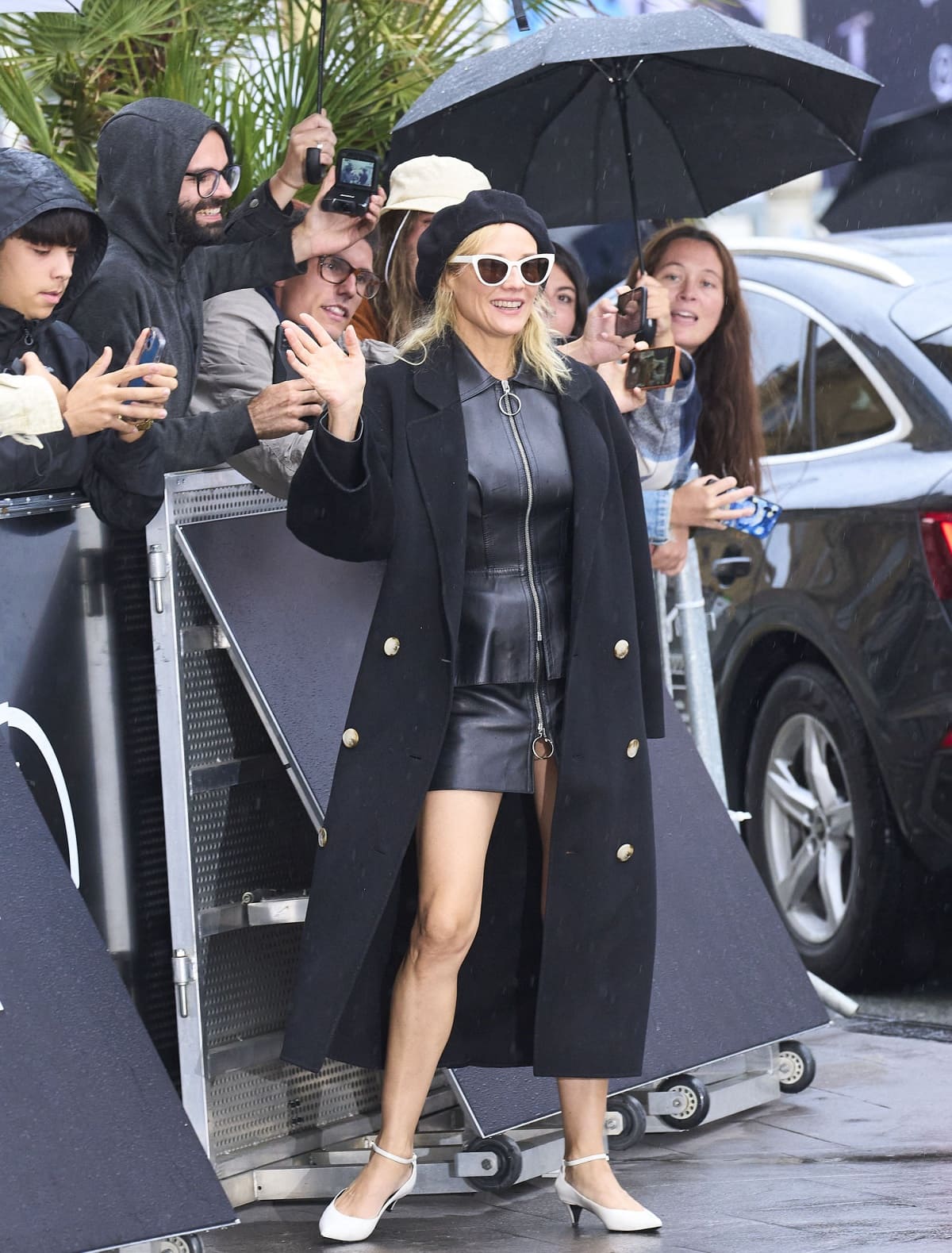 Diane Kruger posing with fans in a black leather dress, a beret, cat-eye sunglasses, and white ankle-strap pumps
