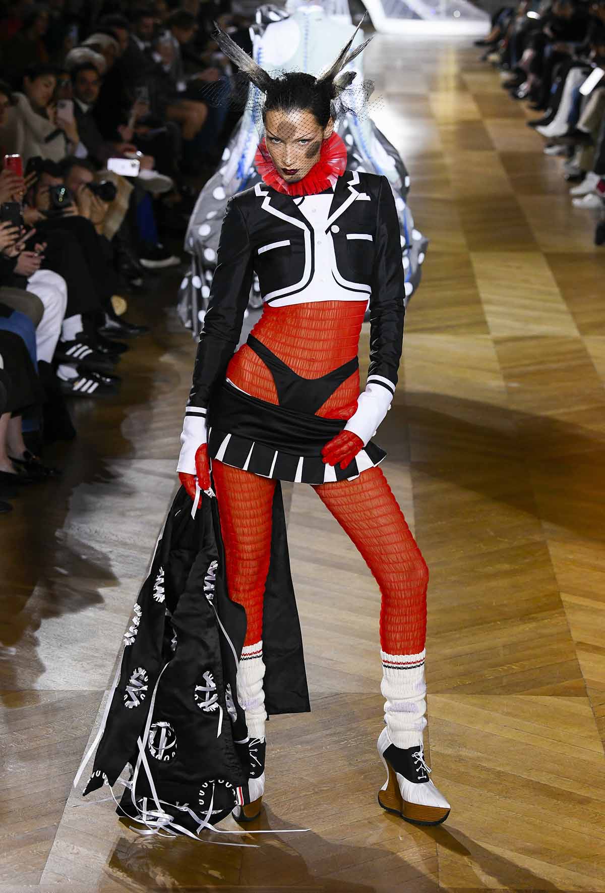 Bella Hadid looks cartoonish in her Thom Browne ensemble, featuring a shitted red jumpsuit, black mini skirt, crop tuxedo, and black underwear