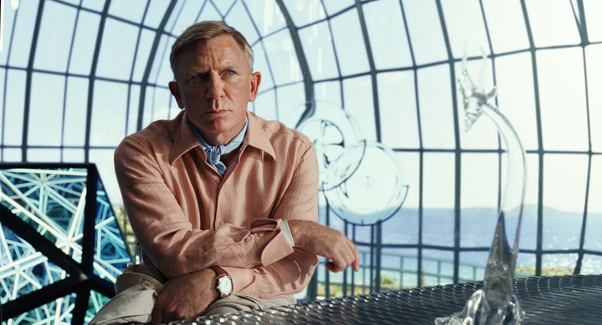 Daniel Craig, as detective Benoit Blanc, appears deep in thought in Glass Onion: A Knives Out Mystery, sitting in an intricately designed glass room overlooking the sea