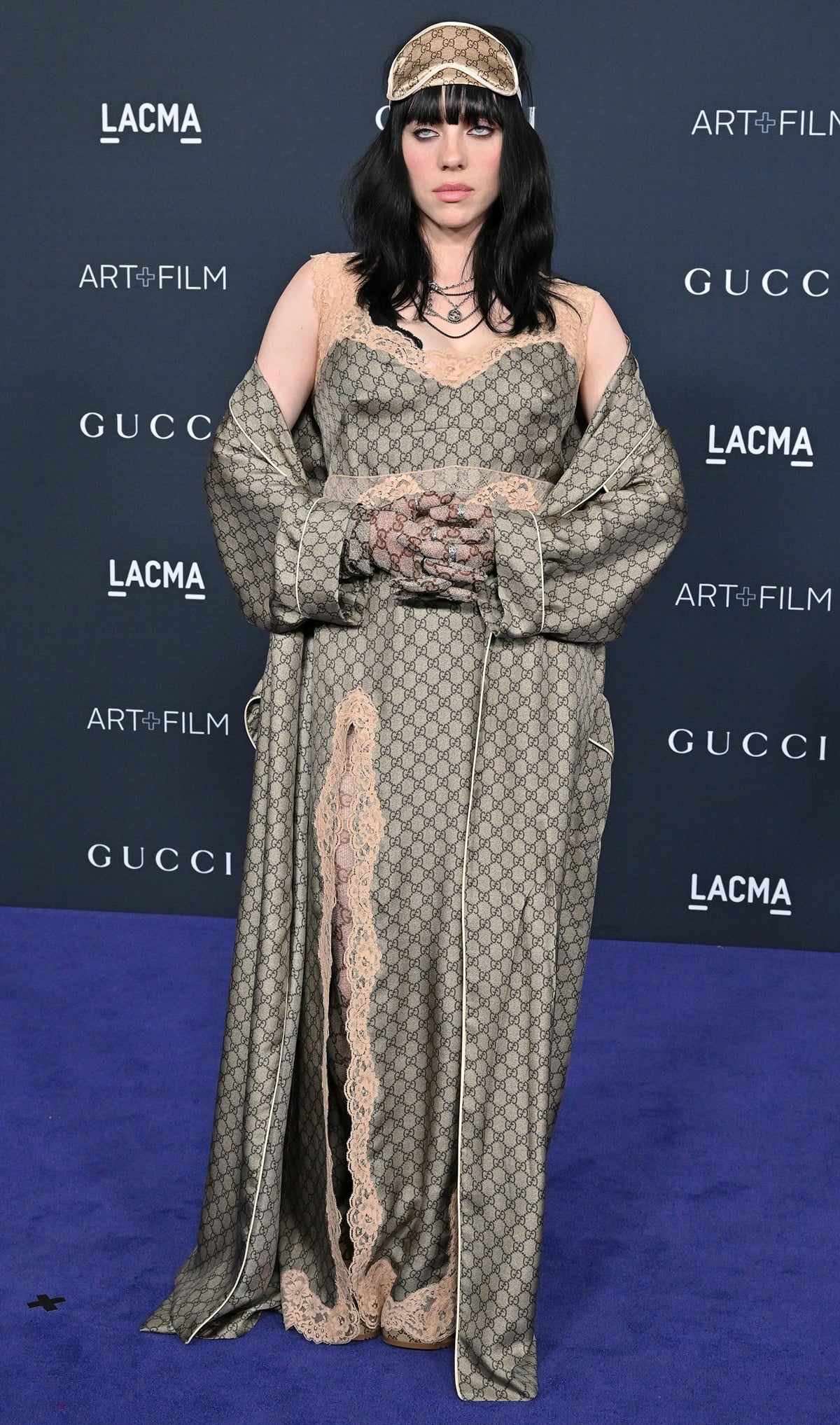 Billie Eilish in Gucci monogrammed form-fitting slip dress paired with a floor-length silk robe, GG tights, gloves, and a sleep mask