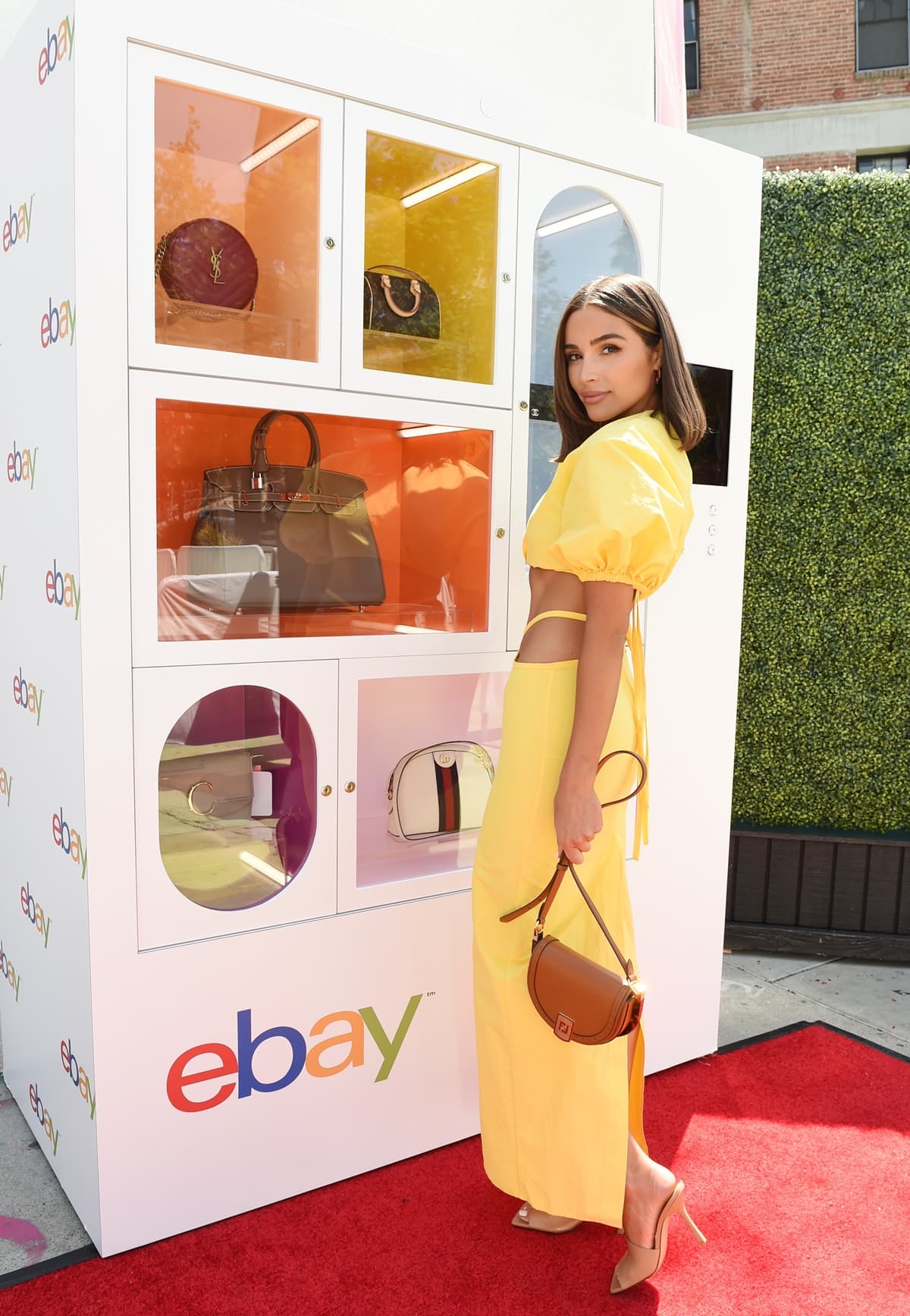 Olivia Culpo, in an outfit by Christopher Esber with Bottega Veneta sandals and a Fendi Moonlight saddle crossbody bag, poses in front of eBay's luxury handbag machine
