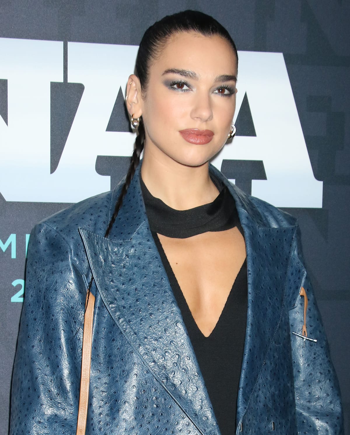 Dua Lipa pulls her raven hair back into a braided ponytail and wears glittery smokey eyeshadow that complements her coat