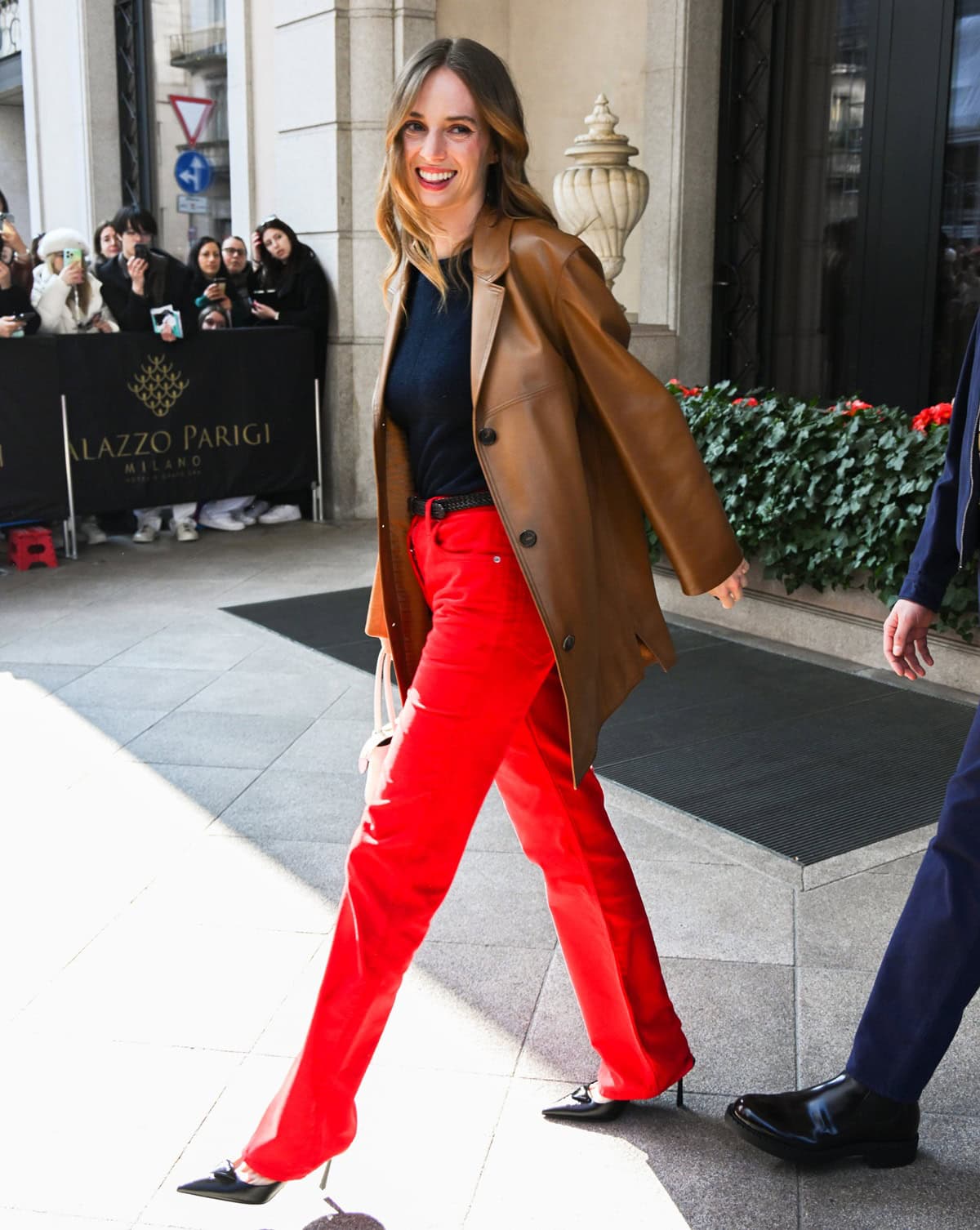 Maya Hawke added a sharp edge to her look with pointed black stiletto heels, pairing them with fire-engine red trousers, a navy knit top, and a caramel leather coat while attending Prada’s Fall/Winter 2025/2026 show at Milan Fashion Week
