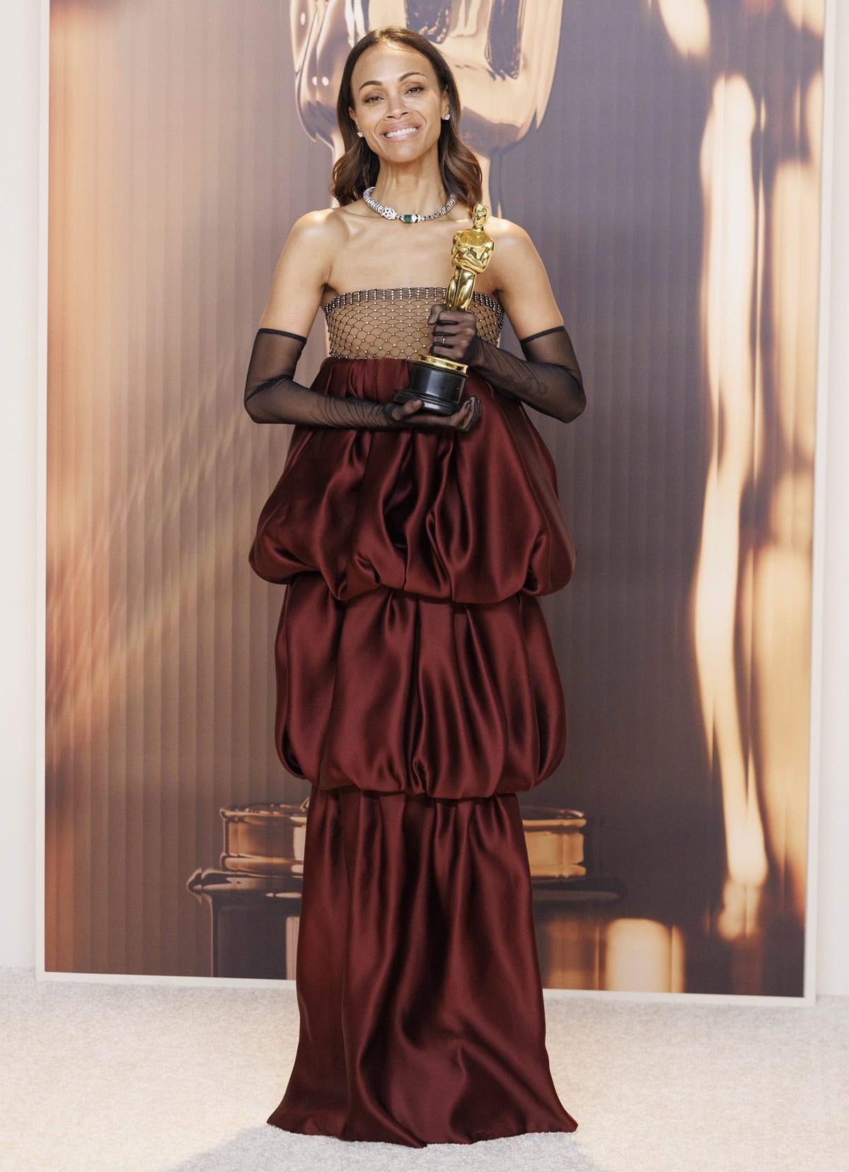 Zoe Saldaña stuns at the 2025 Oscars in a custom Saint Laurent gown featuring a rhinestone mesh bodice, dramatic burgundy bubble hem, and sheer opera gloves