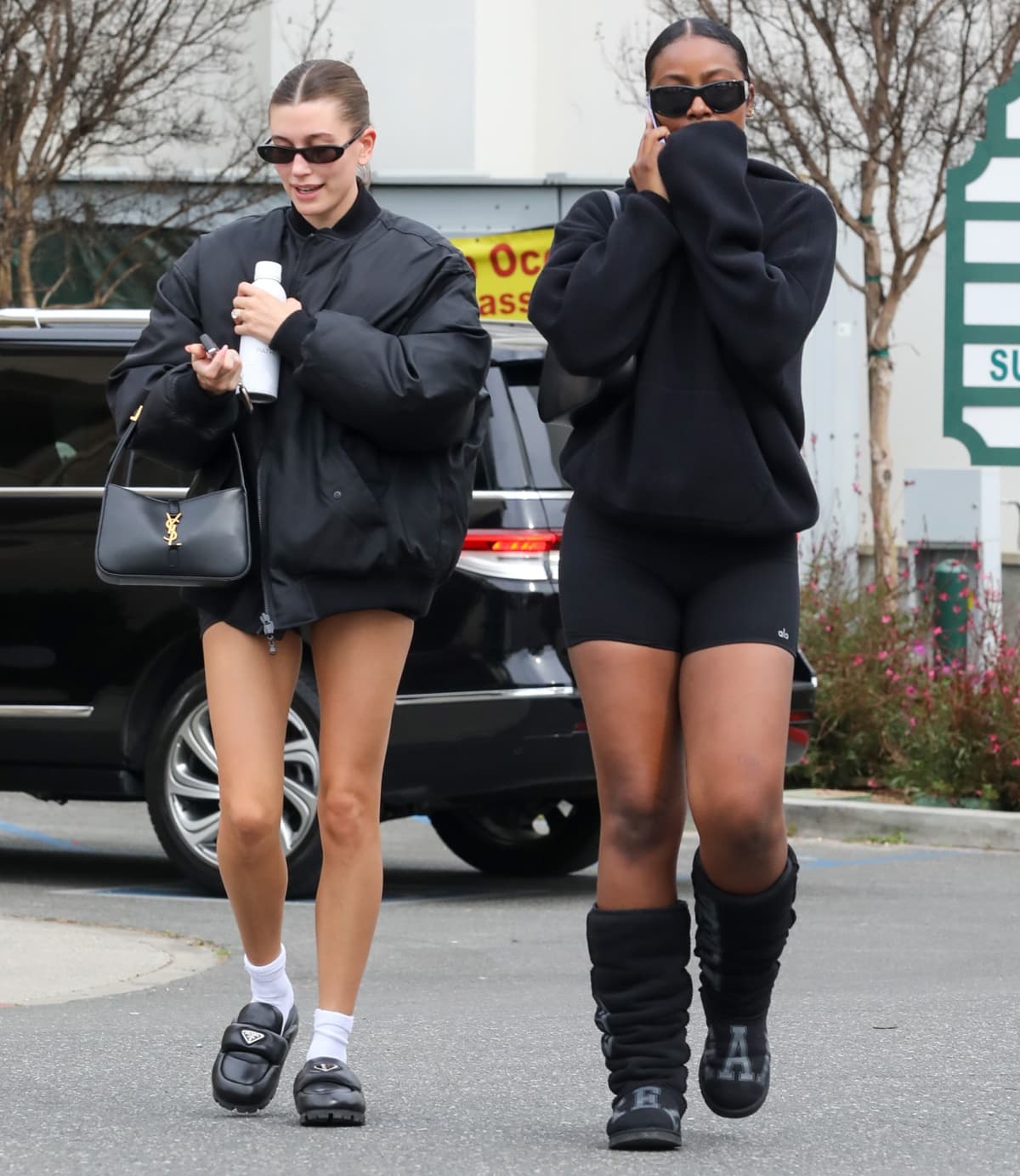 Joined by Justin Skye, Hailey Baldwin wore an oversized black bomber-style puffer coat over tiny black shorts on February 18, 2023, paired with chunky Prada mules, white socks, Matrix-esque sunglasses, a simple shoulder bag, and a white water bottle