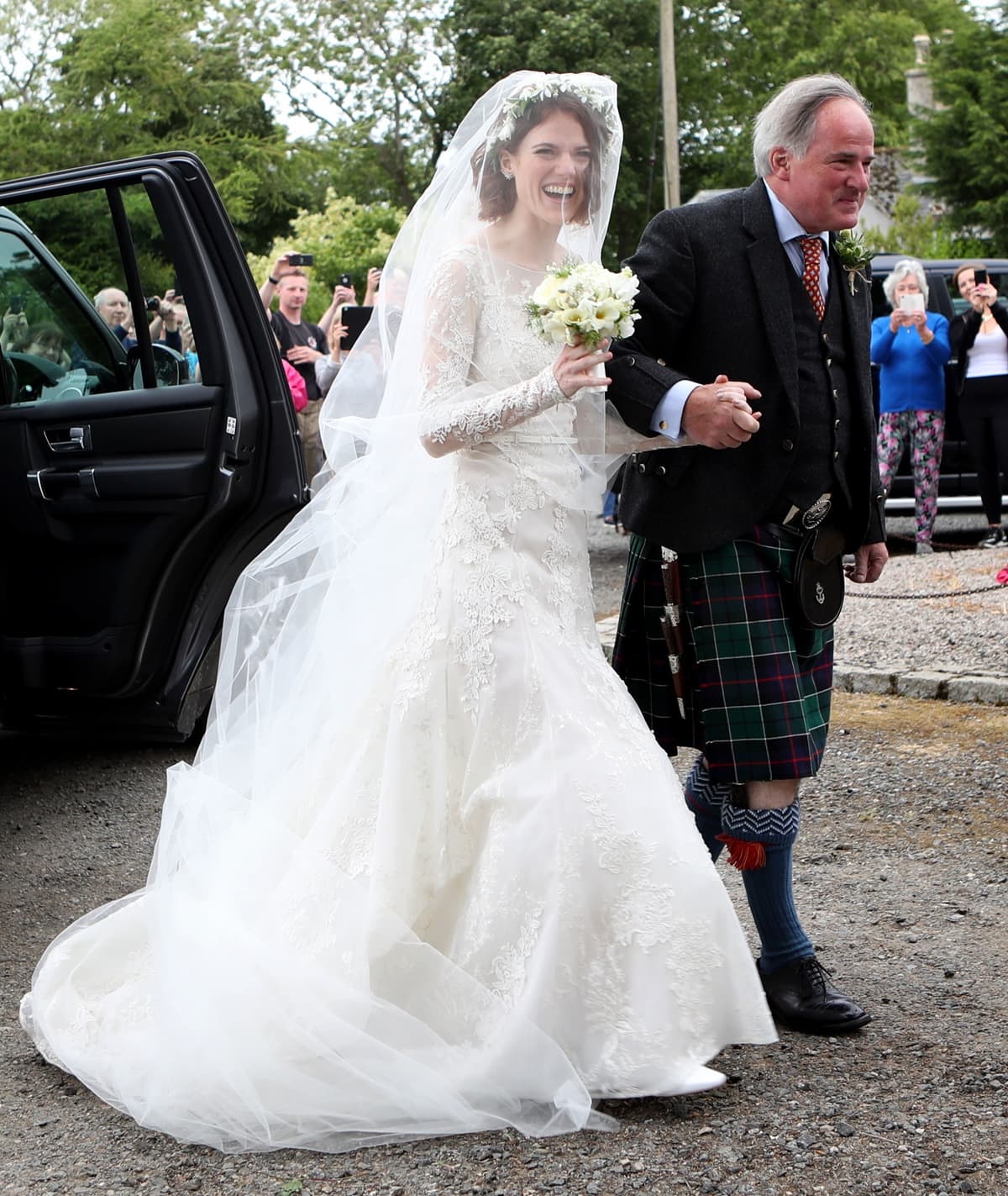 Rose Leslie smiled brightly as she emerged from the car with her father, Sebastian Arbuthnot-Leslie, wearing a vintage-style Elie Saab lace dress featuring a classic fit-and-flare cut, a sweetheart neckline, and long sheer sleeves