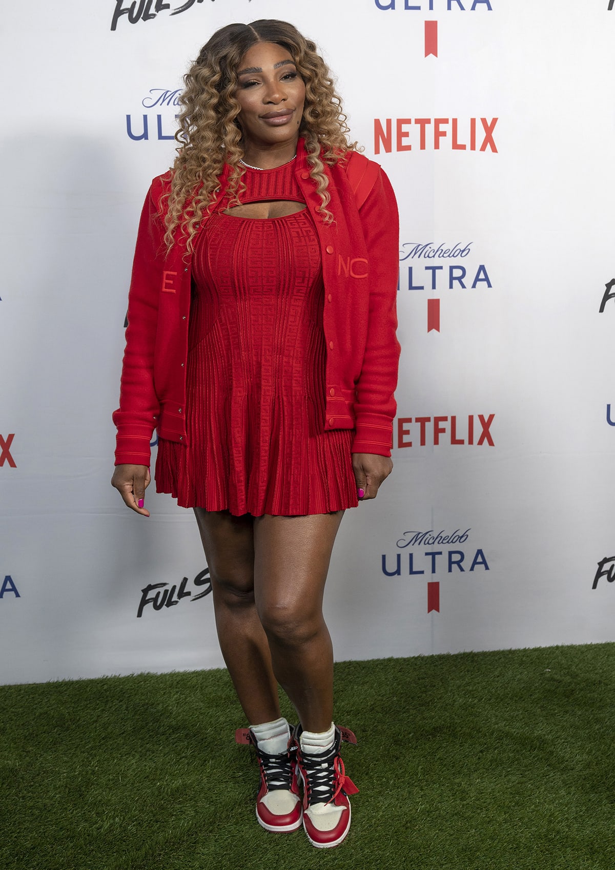 Serena Williams showcases her athletic body in a red minidress with a matching unbuttoned red sweater and white-and-red Jordan high-top sneakers
