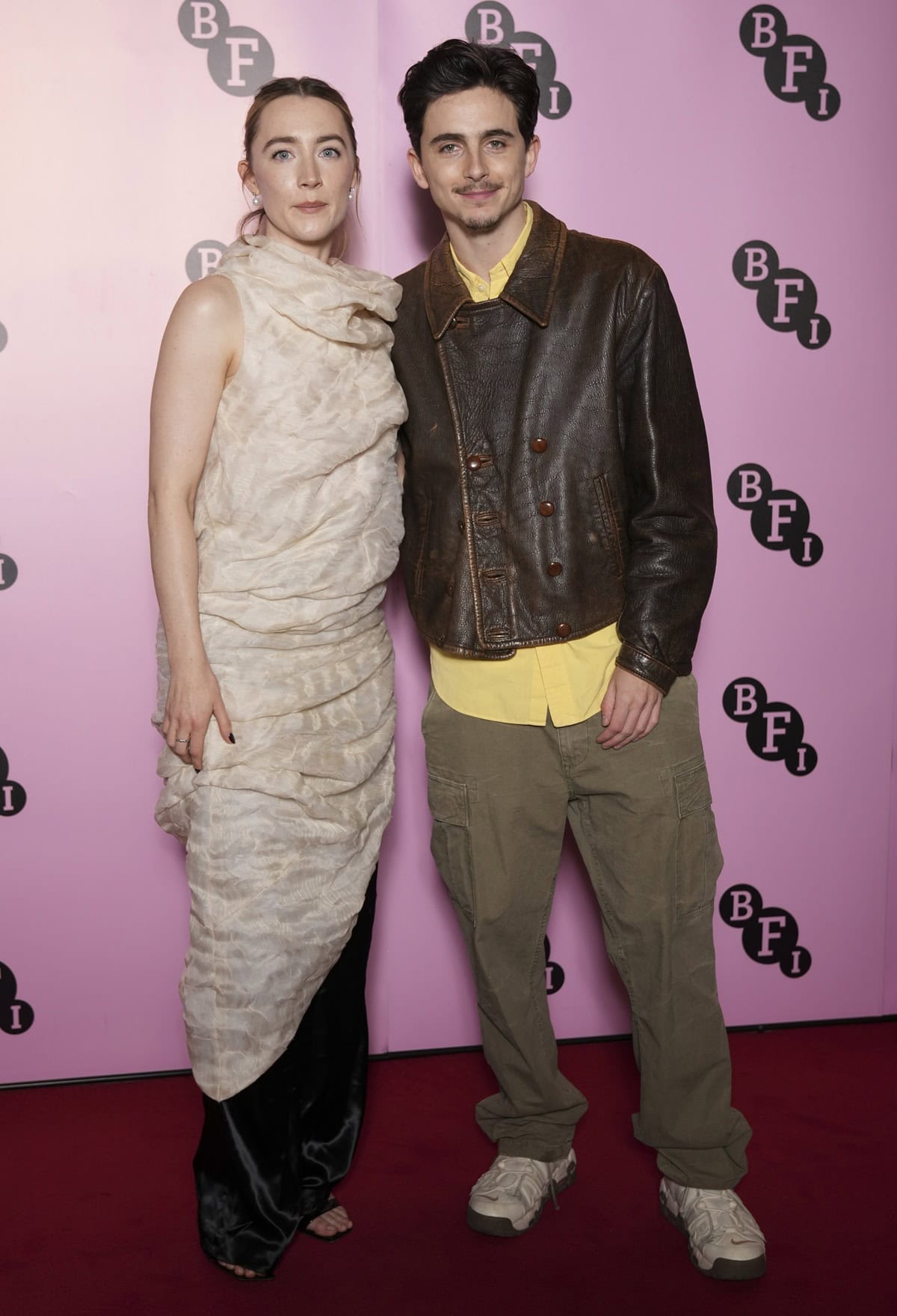 Saoirse Ronan in a draped peachy-cream Khaite organza silk dress with black pants and sandals, alongside Timothée Chalamet in a distressed brown leather jacket, yellow shirt, and cargo trousers at the BFI Southbank In Conversation event in London on December 18, 2024