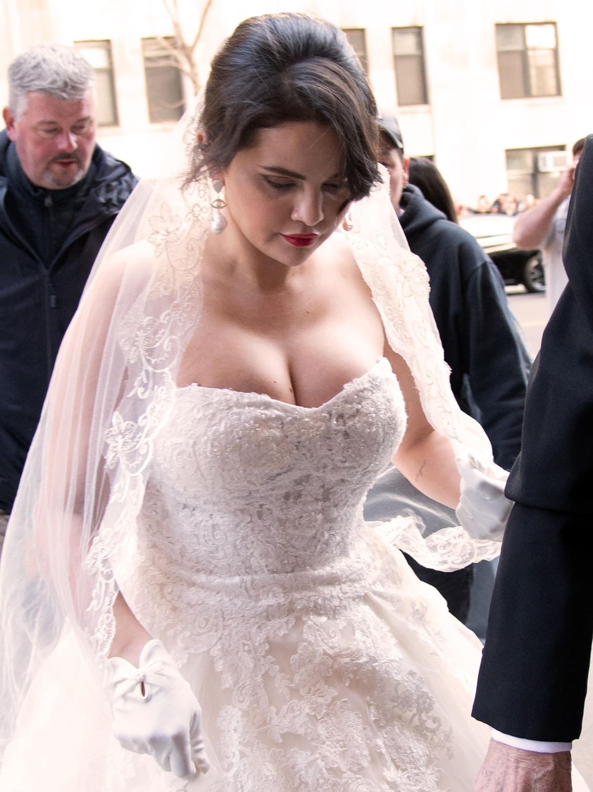 Selena Gomez flaunts cleavage in a beautiful ball gown-style wedding dress with a strapless sweetheart neckline, voluminous skirt, and delicate lace, which she paired with a matching floor-length lace veil, white gloves, pearl drop earrings, and a side-parted updo, completed with a cherry red lip