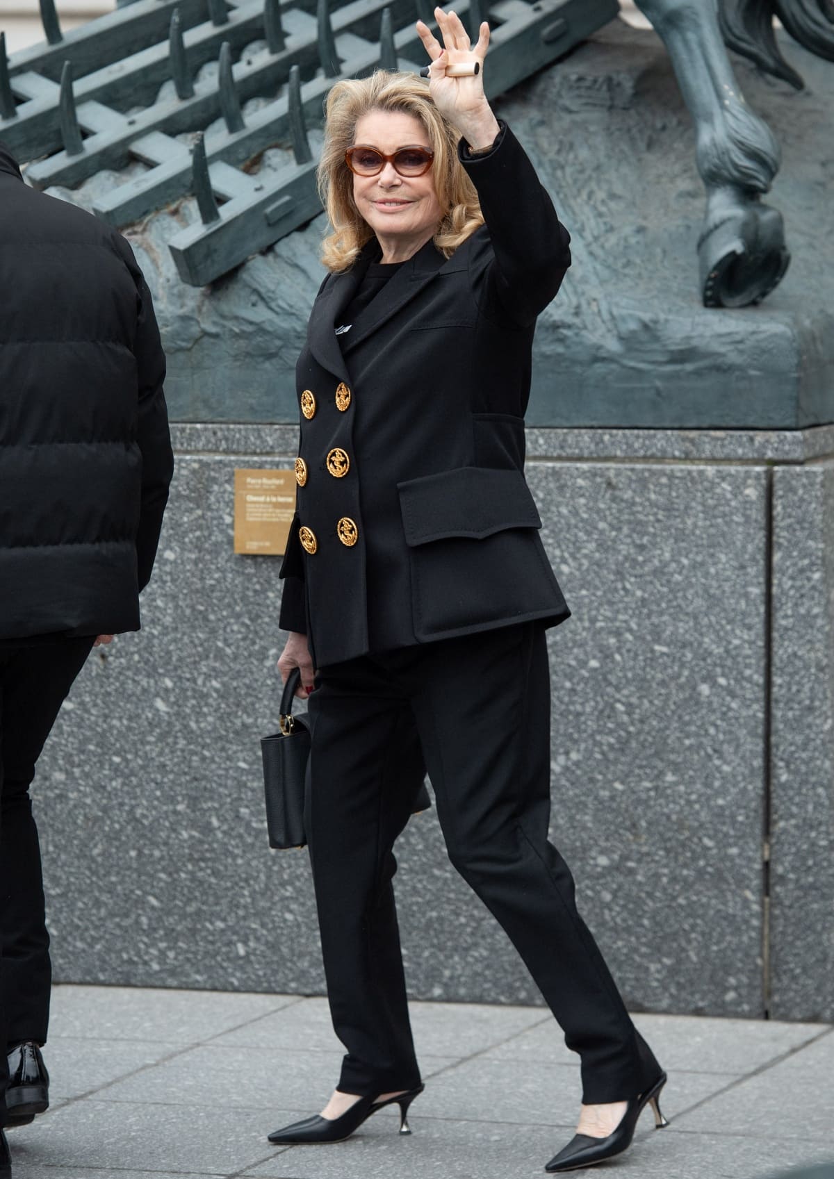Catherine Deneuve exudes timeless Parisian elegance in a black tailored pantsuit with oversized gold buttons, paired with sleek black kitten heels at the Louis Vuitton Fall/Winter 2023 show during Paris Fashion Week on March 6, 2023