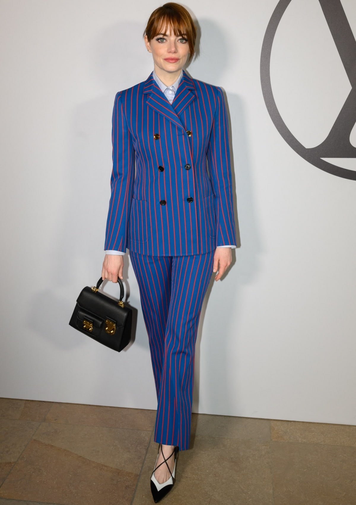 Emma Stone posing in a blue-and-orange pinstriped suit with pointy-toe heels and a black leather top-handle bag