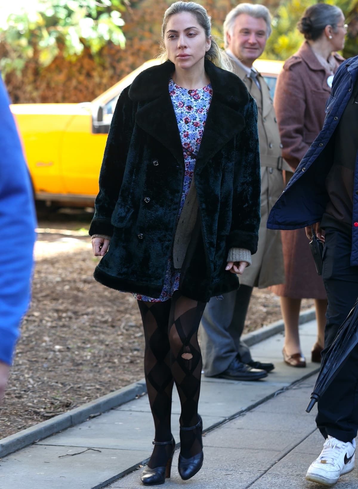 Lady Gaga wearing a crushed blue velvet coat over a floral-printed dress with black harlequin stockings and pointy-toe heels while filming Joker: Folie a Deux