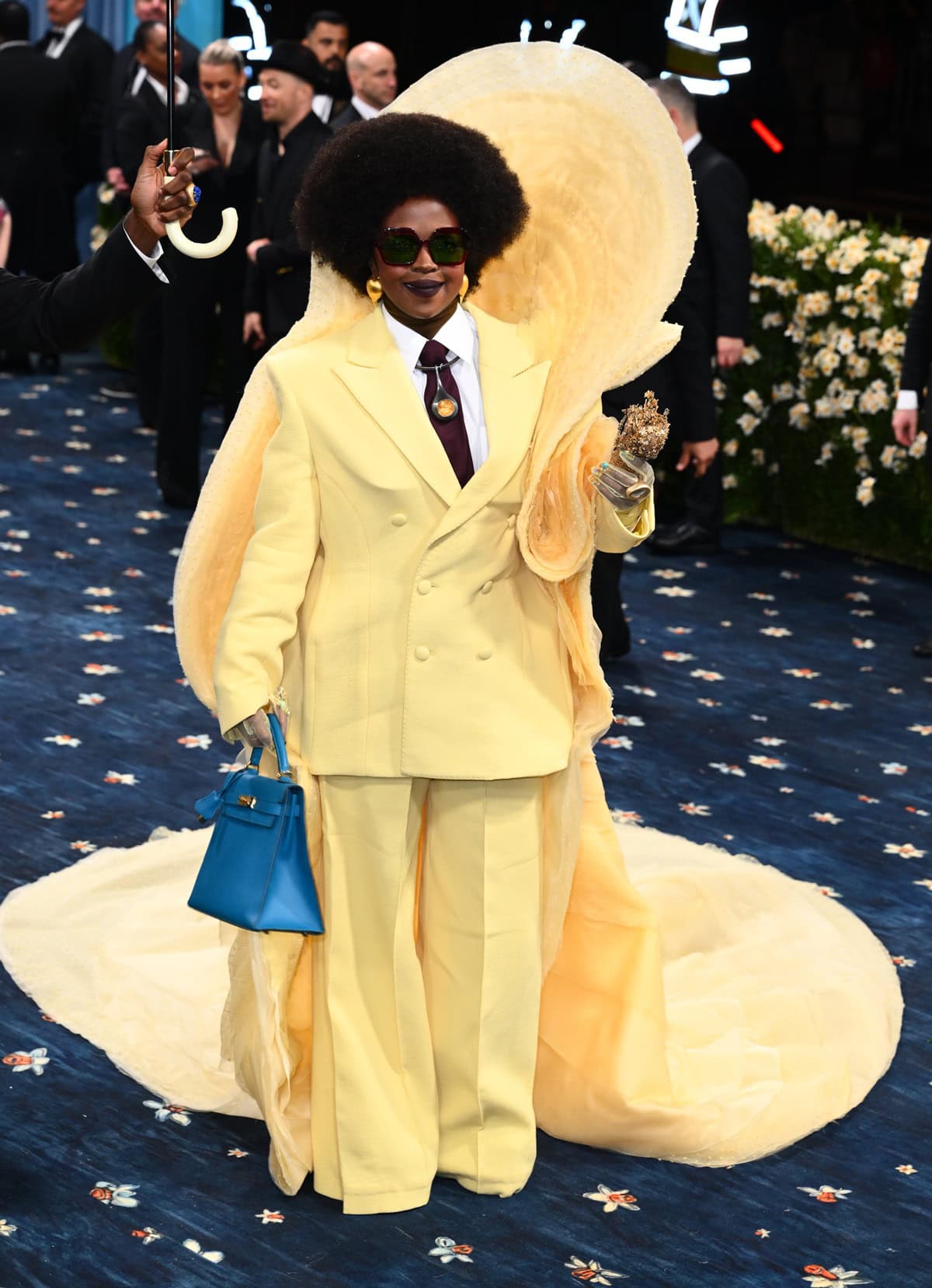 Lauryn Hill made a powerful Met Gala debut in a sunshine-yellow Cheney Chan Couture suit with sculptural cape detailing, pairing the bold ensemble with hidden platform heels and a striking blue Hermès Kelly bag