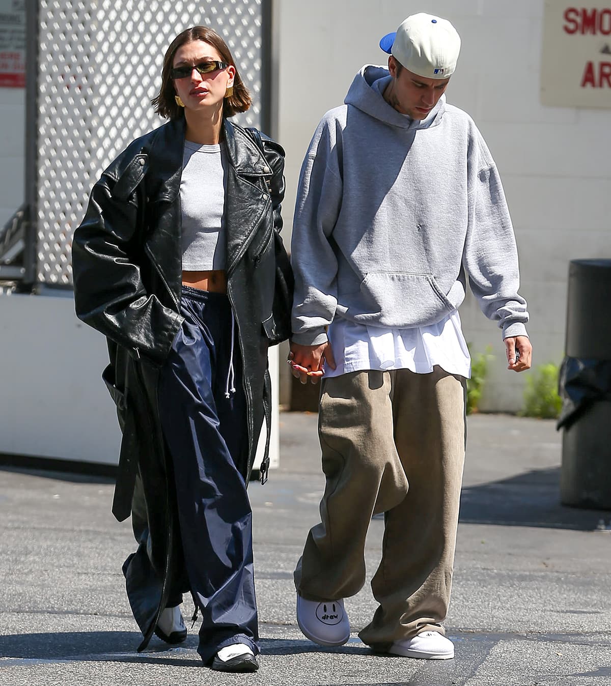 Hailey Bieber flashes her abs in a gray crop tee, blue parachute pants, and maxi-length Balenciaga trench coat, while Justin Bieber looks cool in a gray hoodie, taupe pants, and House of Drew shoes