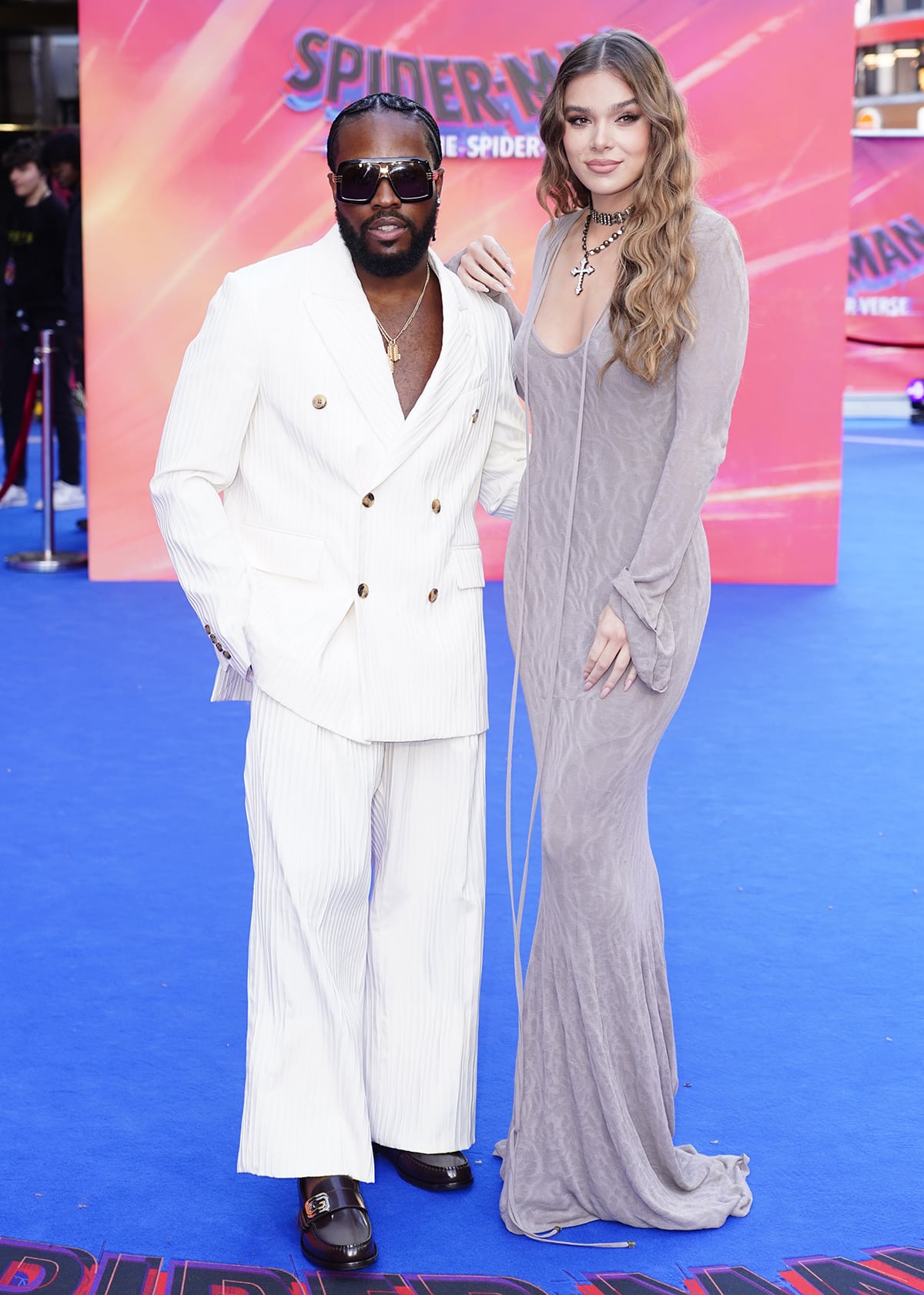 Hailee Steinfeld towers over Shameik Moore, who looks dapper in a white suit, layered gold necklaces, and black sunglasses