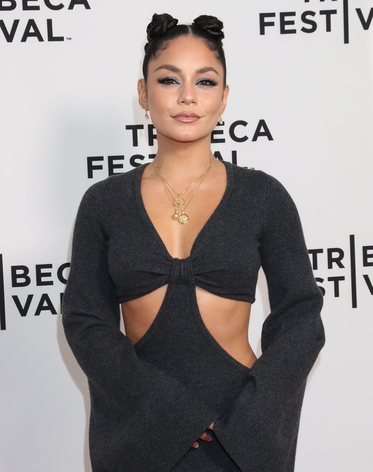 Vanessa Hudgens styles her dark hair in sleek space buns with swirled baby hairs and wears fake eyelashes, smokey blue-gray eyeshadow, and nude lipstick