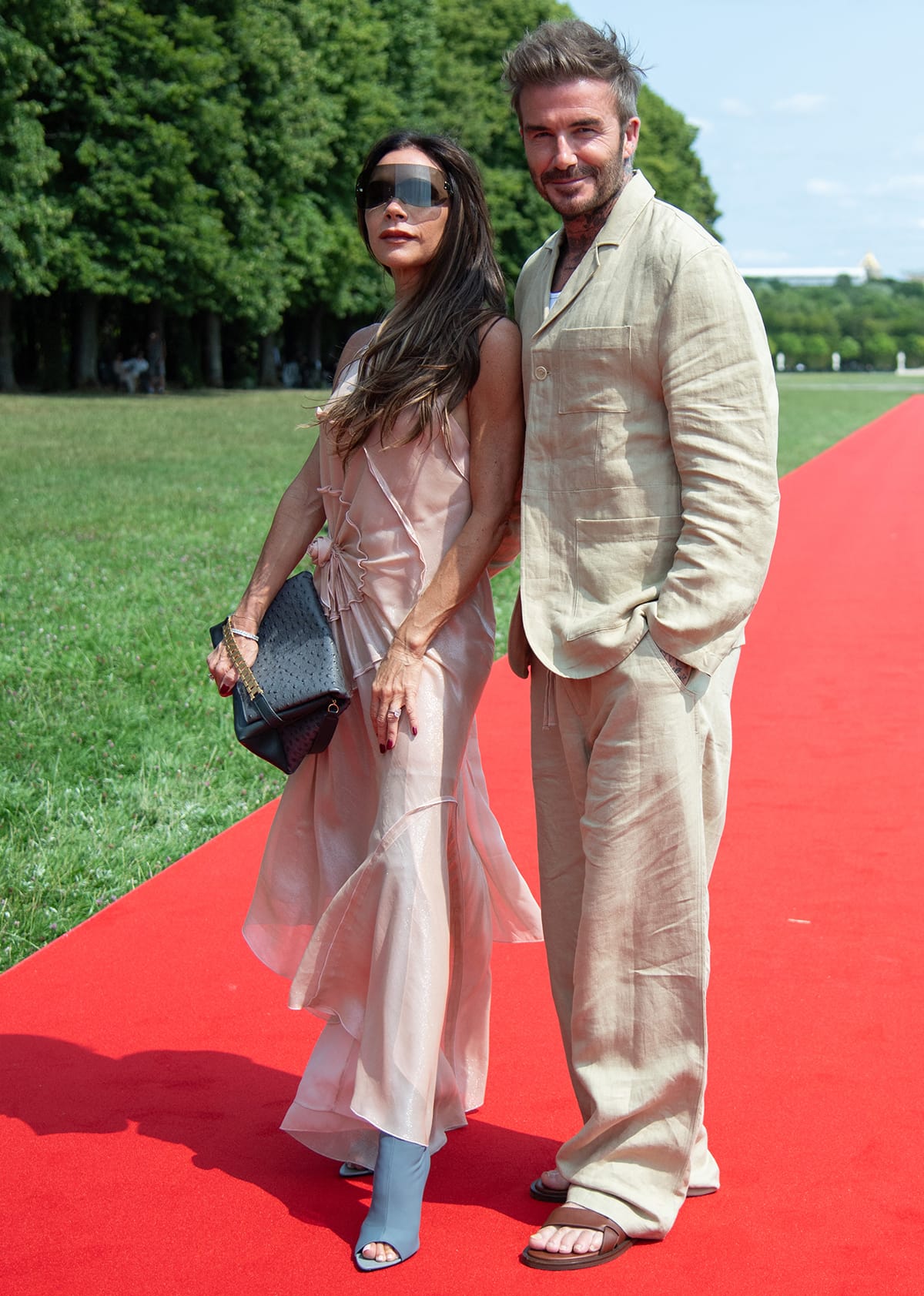 Victoria Beckham and David Beckham pose on the red carpet in matching neutrals, with the athlete in a beige suit and brown sandals and the pop star in a pink slip dress and thigh-high boots