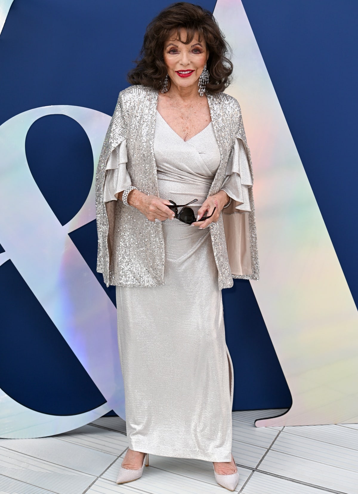 Dame Joan Collins wearing a champagne gold sequin gown with a shimmering cape and a side slit
