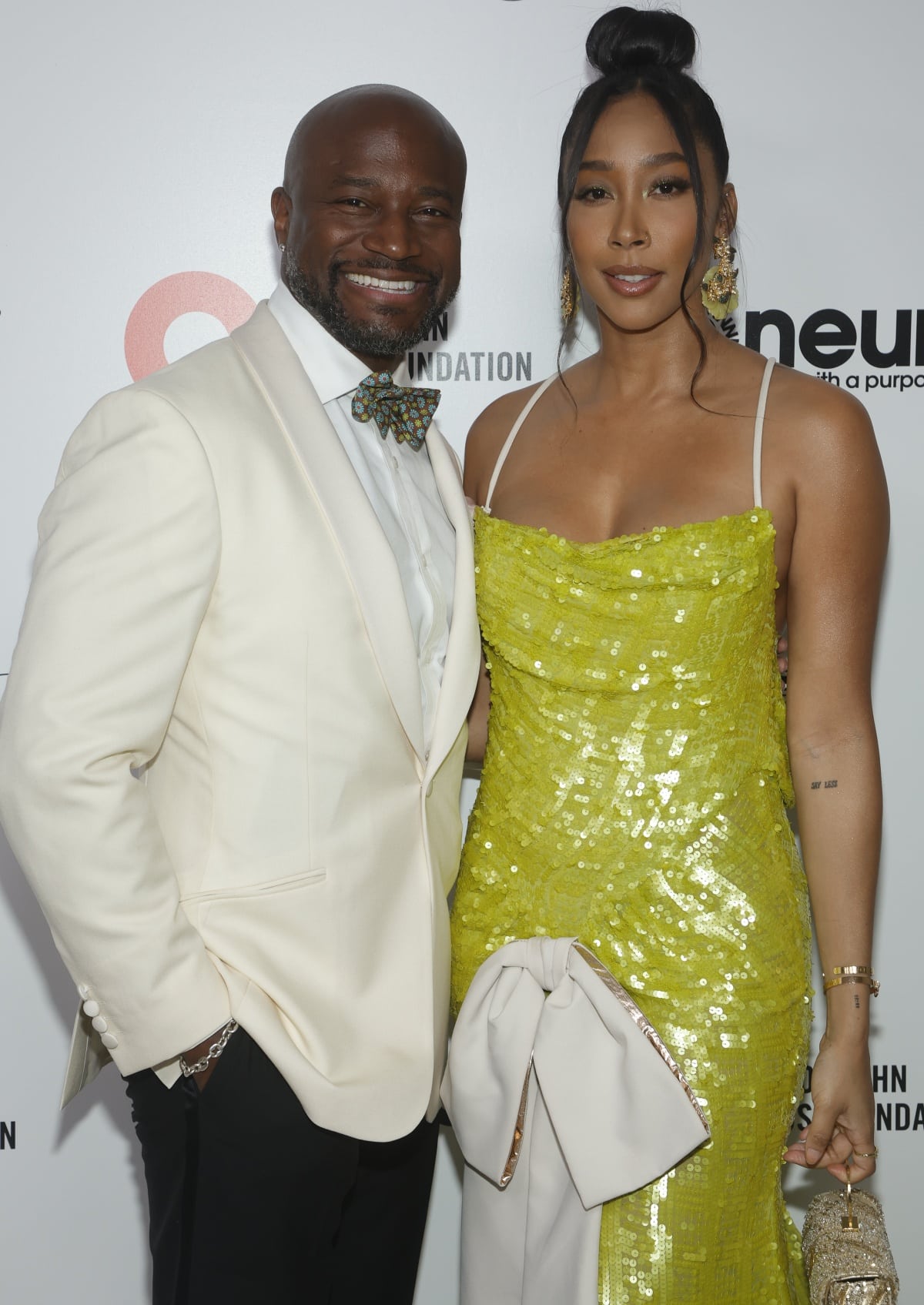 Taye Diggs in a white suit with a printed bowtie and Apryl Jones in an embellished gown with bow detail at the 31st Annual Elton John AIDS Foundation Academy Awards Viewing Party