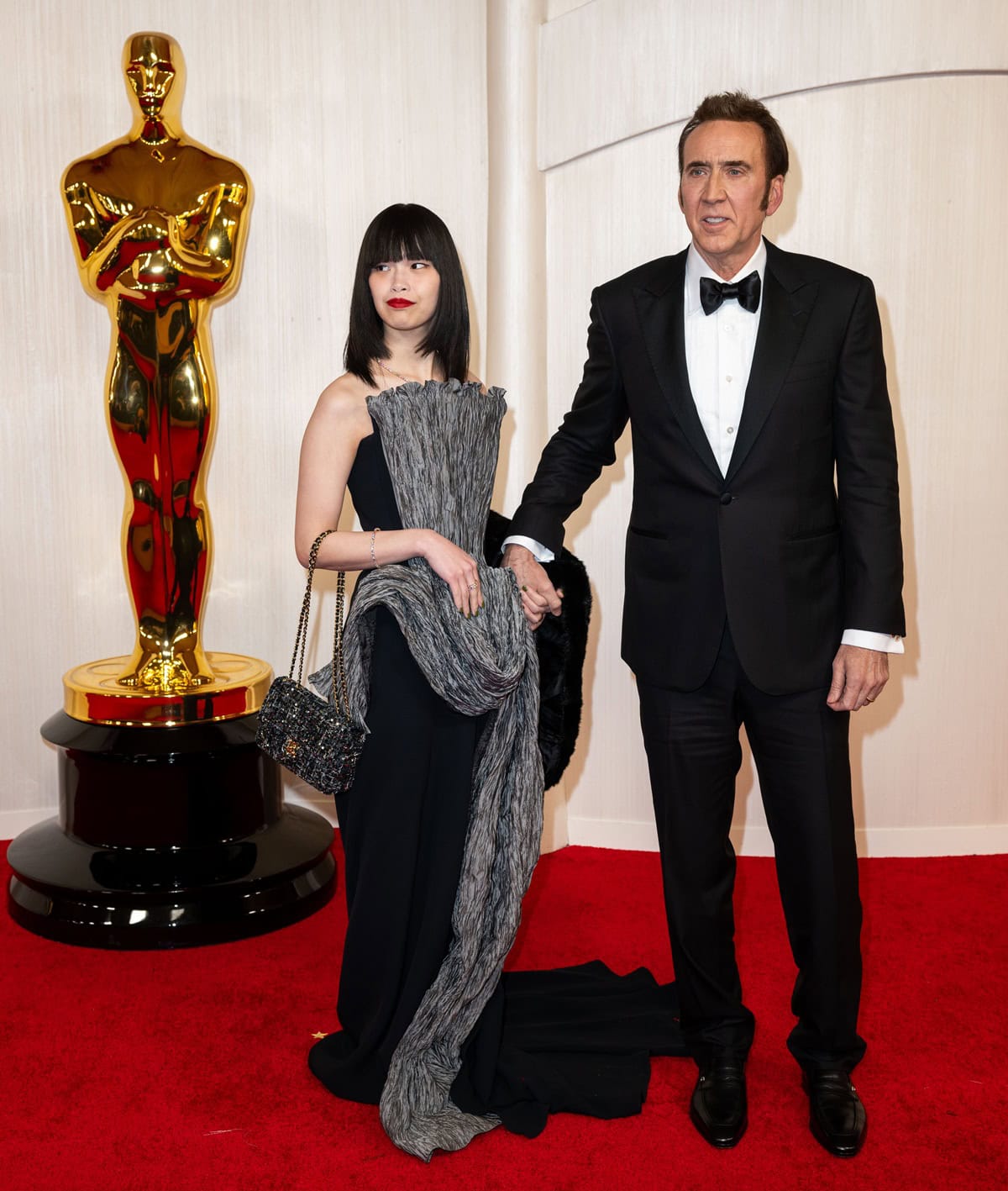 Nicolas Cage and Riko Shibata made a striking pair at the 96th Oscars, coordinating in black and gray — Cage in a classic tuxedo and Shibata in a sculptural floor-length gown featuring textured plissé detailing and a gem-embellished handbag