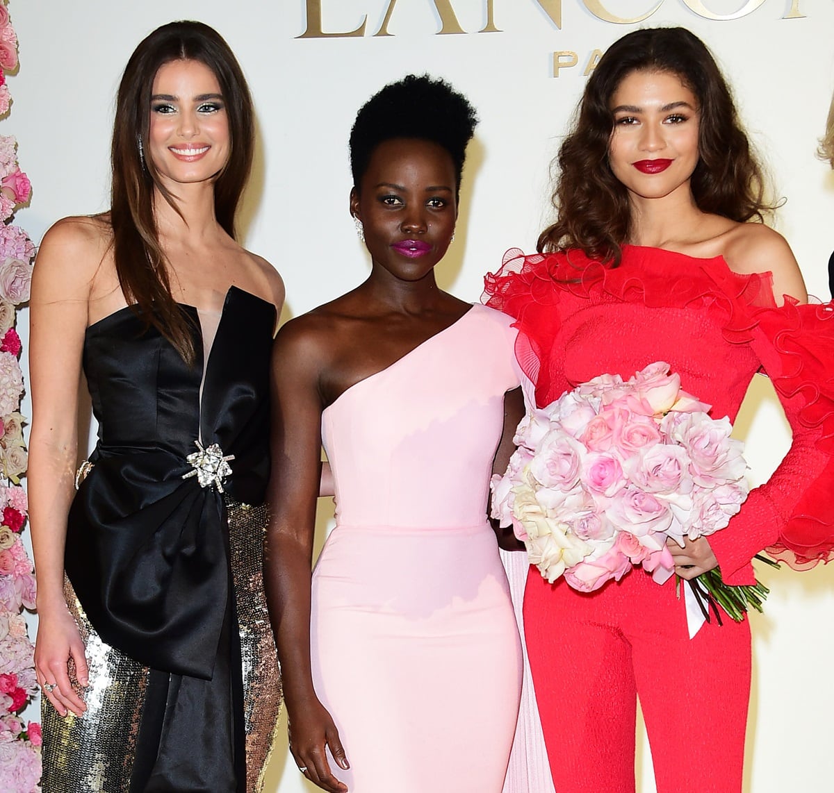 Lupita Nyong'o, looking radiant in a soft pink one-shoulder gown, stands alongside Zendaya and Taylor Marie Hill, embracing her elegance as the shortest among the trio