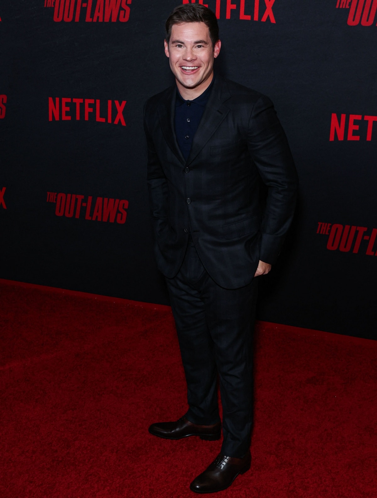 Adam DeVine looks sharp in a dark, tailored suit paired with polished brown dress shoes at the premiere of The Out-Laws, combining classic style with a modern touch