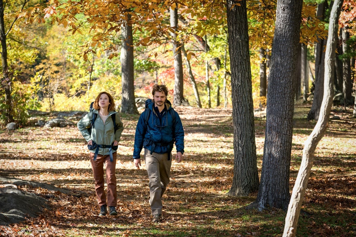 Ellie Kemper and Luke Grimes embrace the rustic spirit of the wilderness in casual hiking attire, sporting earthy-toned cargo pants, sturdy boots, and layered outdoor jackets while trekking through the autumn foliage in Happiness for Beginners