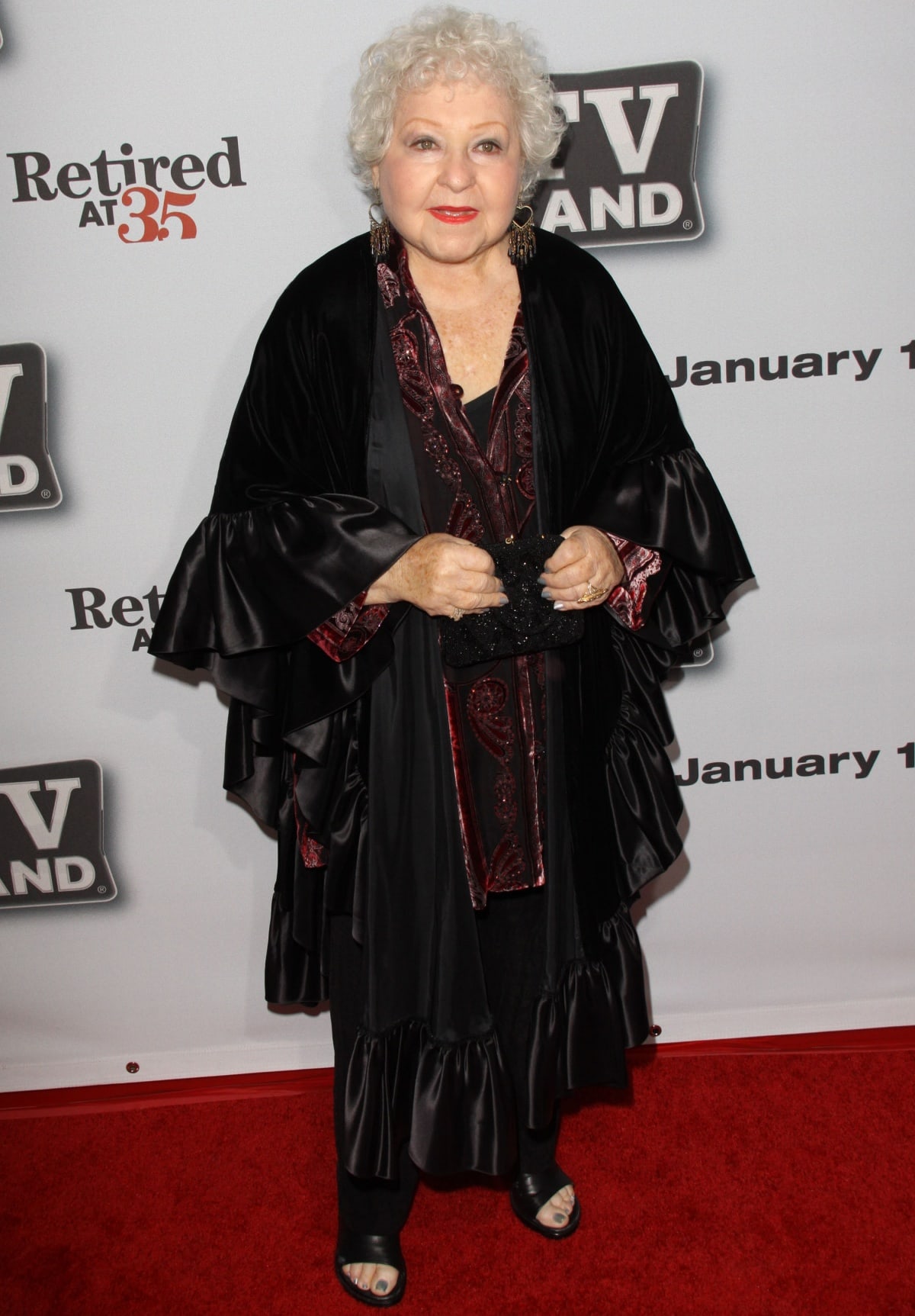Estelle Harris looks elegant in a flowing black ensemble with satin details, pairing it with a rich burgundy blouse and a chic black clutch