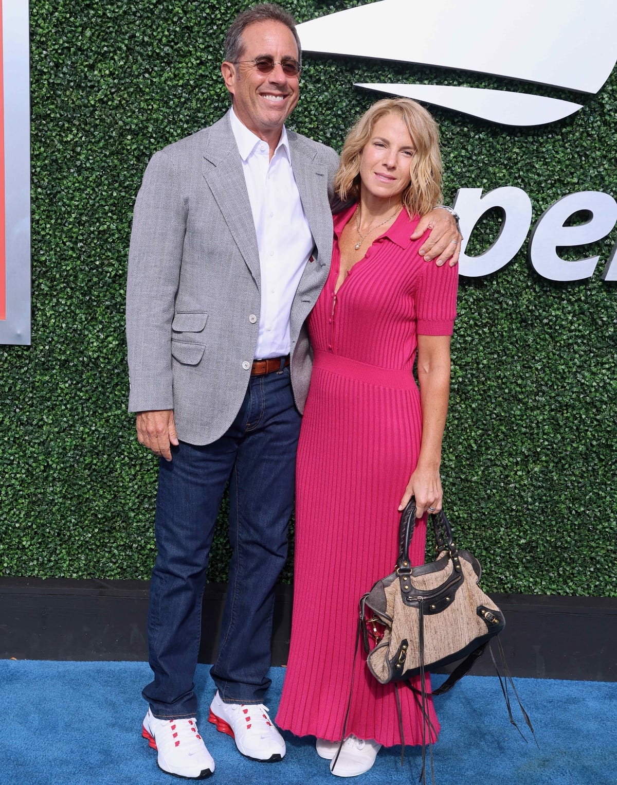 Jerry Seinfeld keeps it casual in a light gray blazer and jeans, while Jessica Seinfeld stuns in a vibrant pink pleated dress paired with a chic handbag, both completing their looks with matching white sneakers