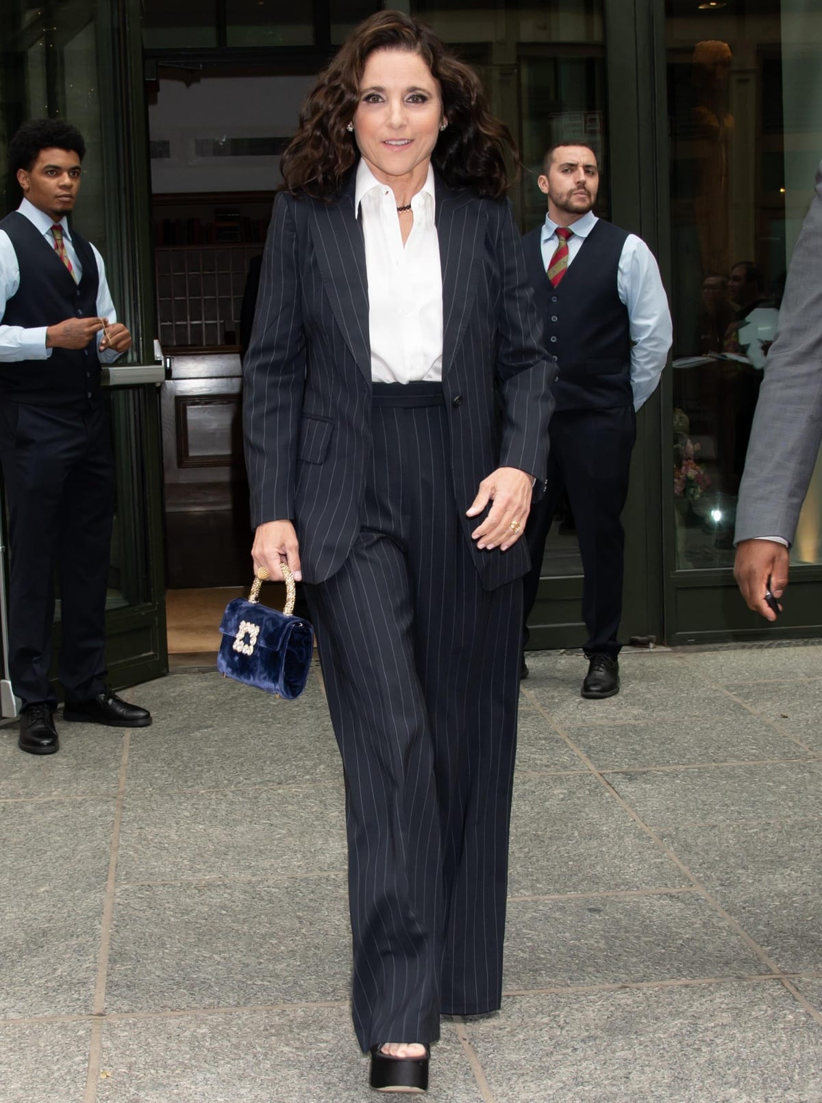 Julia Louis-Dreyfus exudes power and sophistication in a tailored pinstripe suit paired with platform heels and a chic velvet handbag, completing her look with a crisp white shirt