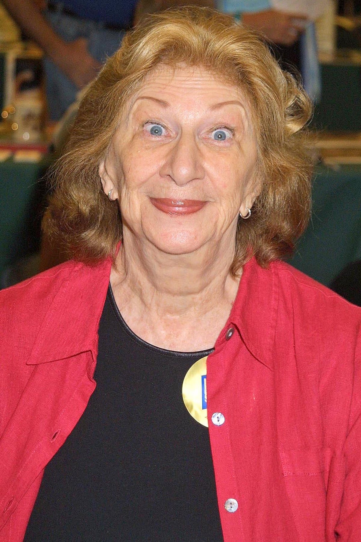 Liz Sheridan smiles brightly in a casual yet bold look, pairing a red button-down shirt with a black top